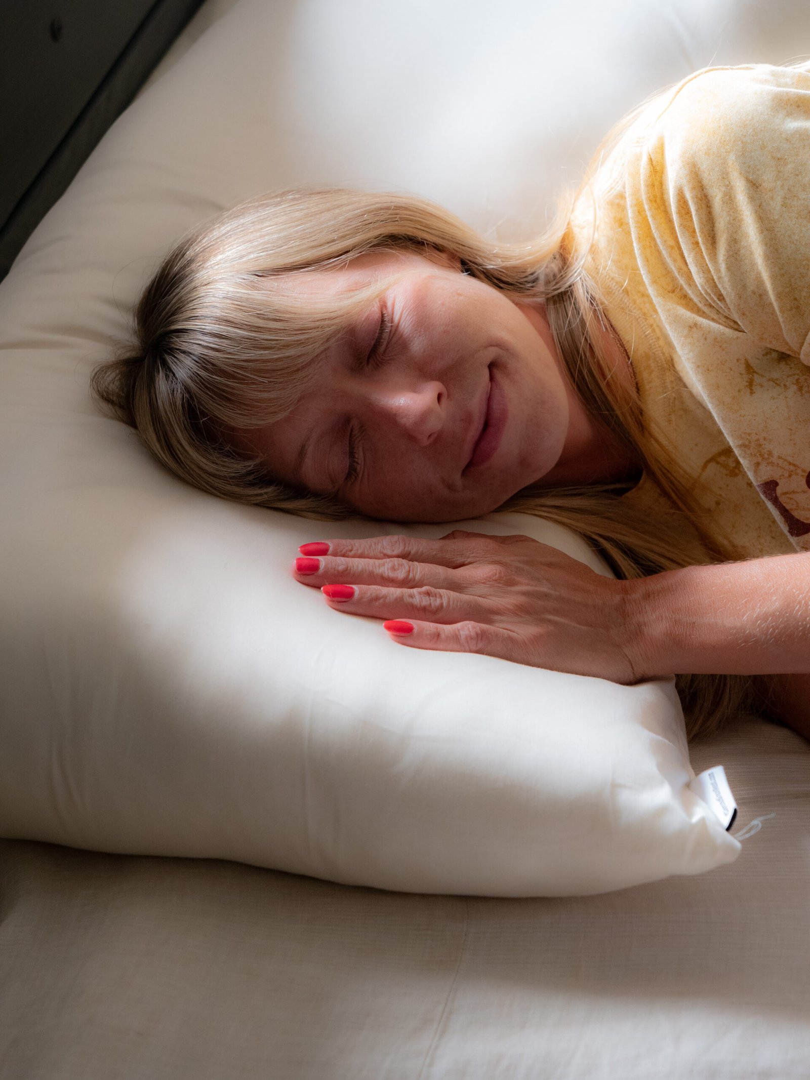 A person with blonde hair lies on a white pillow, eyes closed, wearing a yellow shirt and has red painted nails.