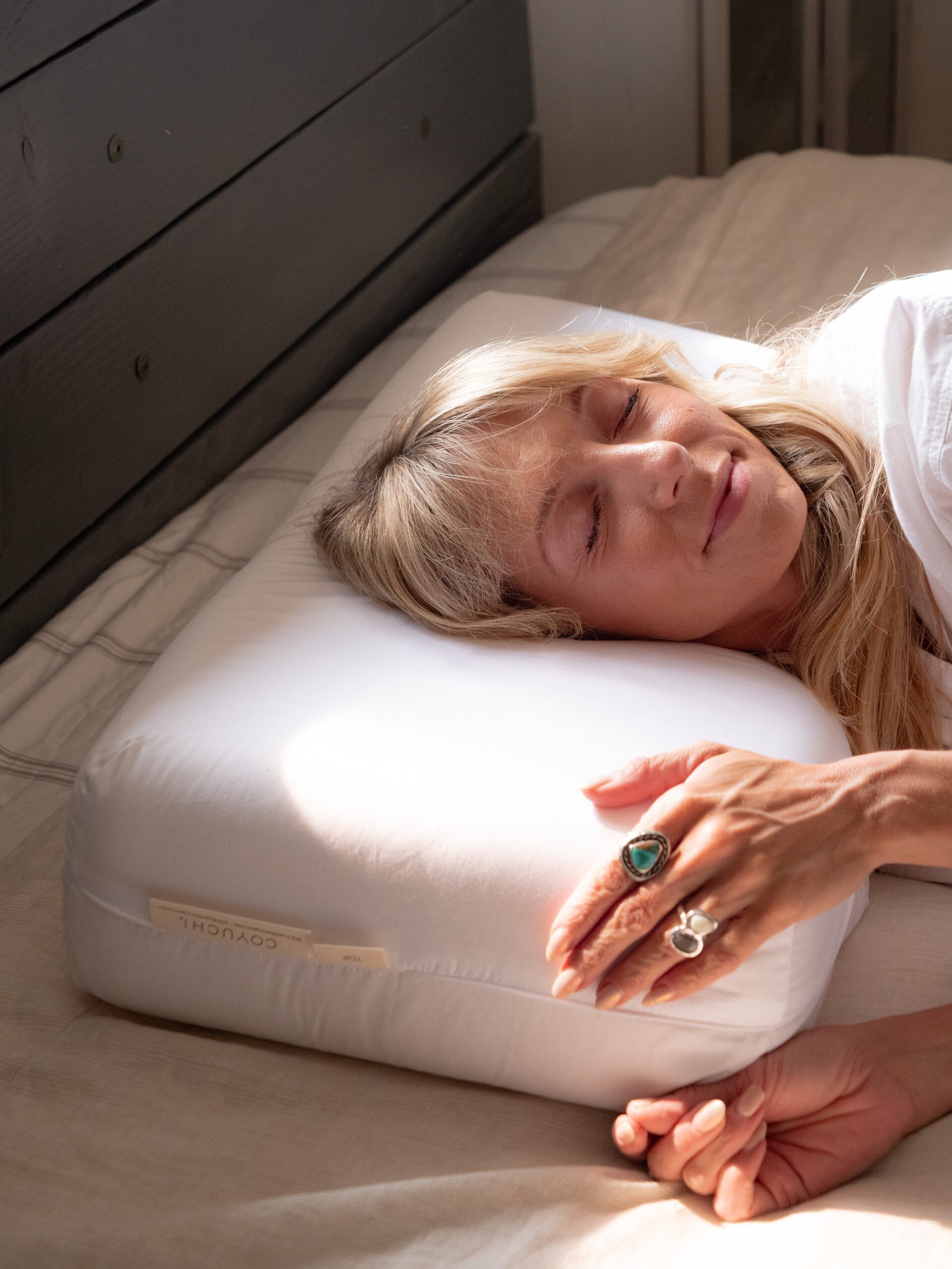 A woman with blonde hair rests on a white pillow, eyes closed, wearing rings and a white shirt, lying on a beige bed.