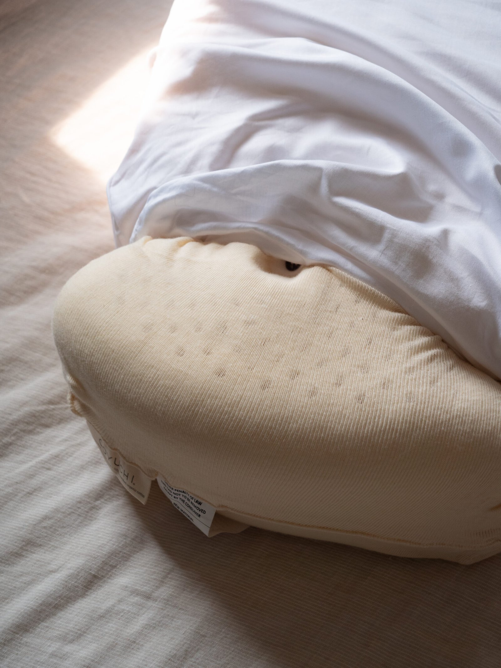 A beige, oval latex pillow partially covered by a white sheet rests on a light-colored bed.