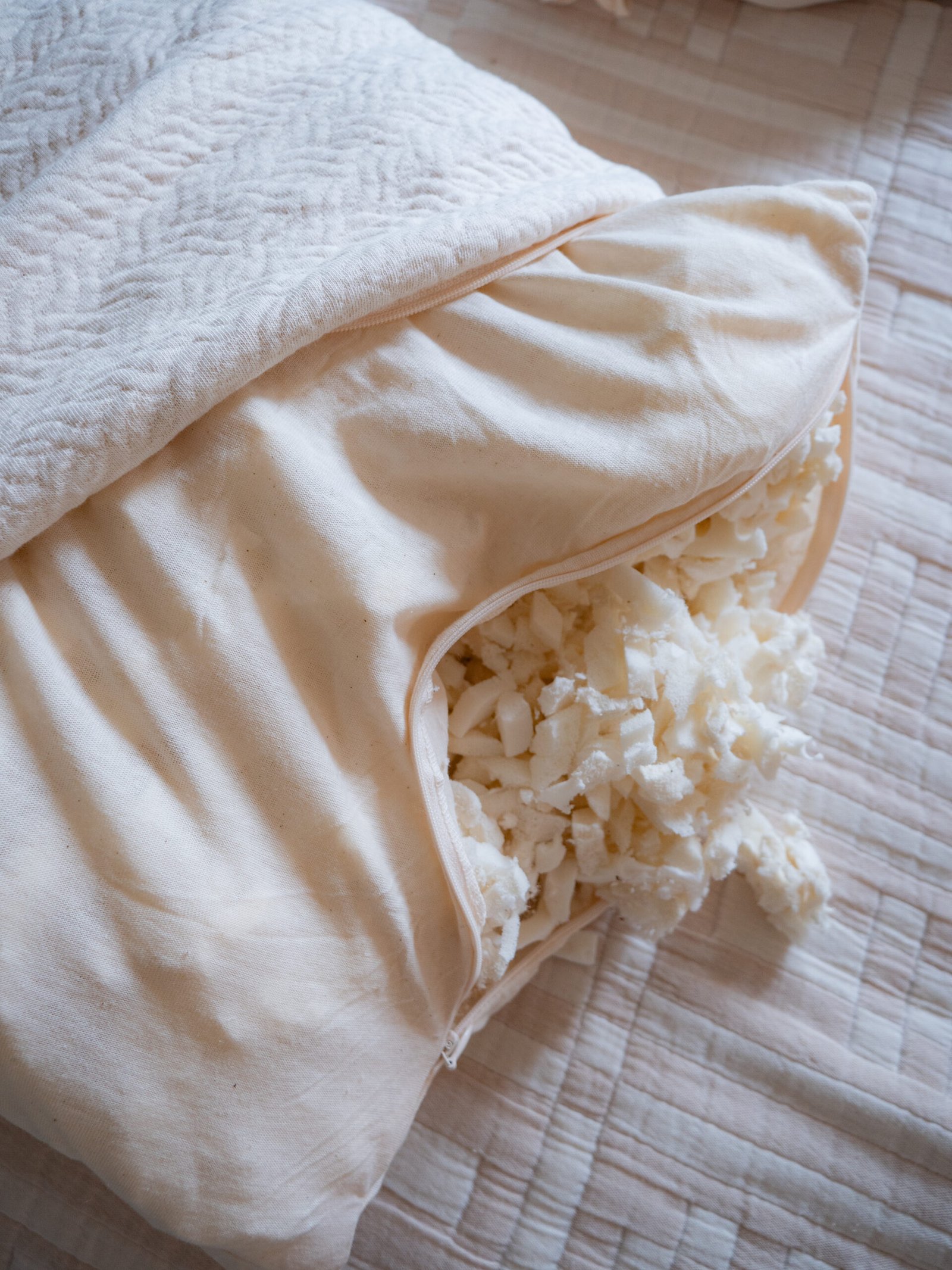 A cream-colored pillow with its zipper open reveals shredded white filling spilling out onto a quilted bedspread.