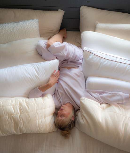 A person in light pajamas is lying in bed surrounded by multiple large white pillows, with one leg bent and arm resting on a pillow.