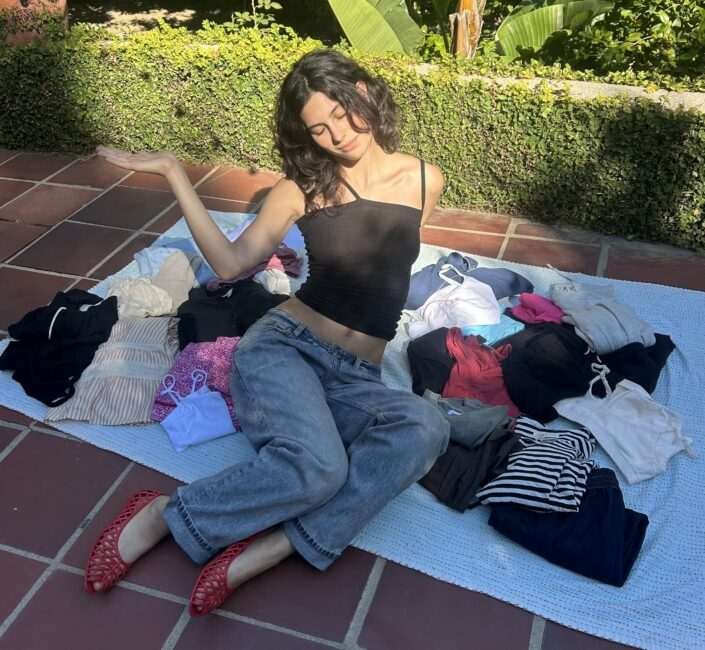 A woman sits on a blanket outdoors surrounded by folded clothes, raising one hand with a nonchalant expression.