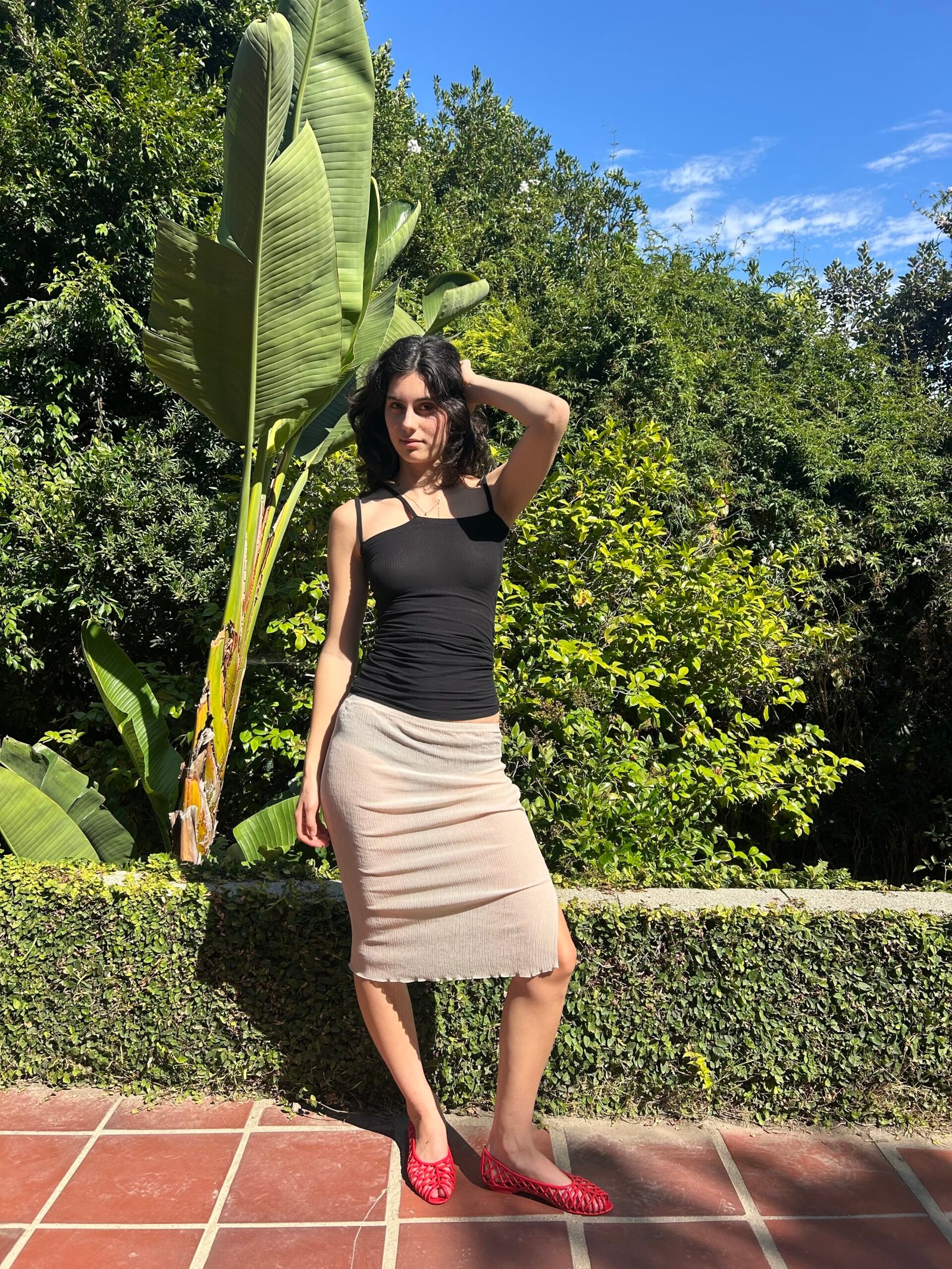 A woman in a black tank top and beige skirt stands on a tiled patio in front of green foliage, with one hand on her head.