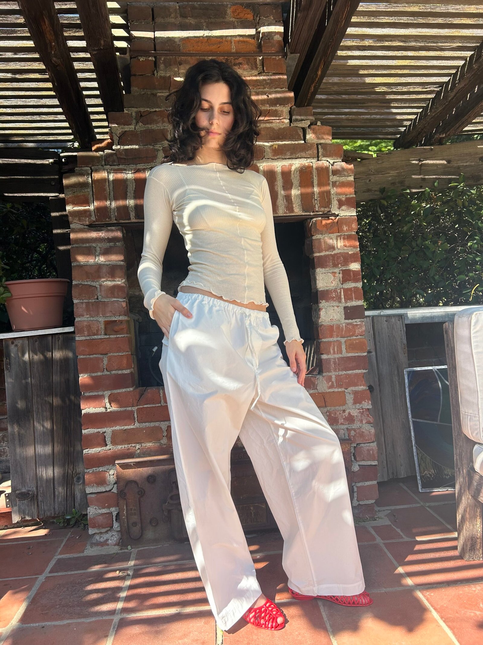A person with shoulder-length dark hair stands in front of a brick outdoor fireplace, wearing a sheer long-sleeve top, loose white pants, and red socks with white spots.