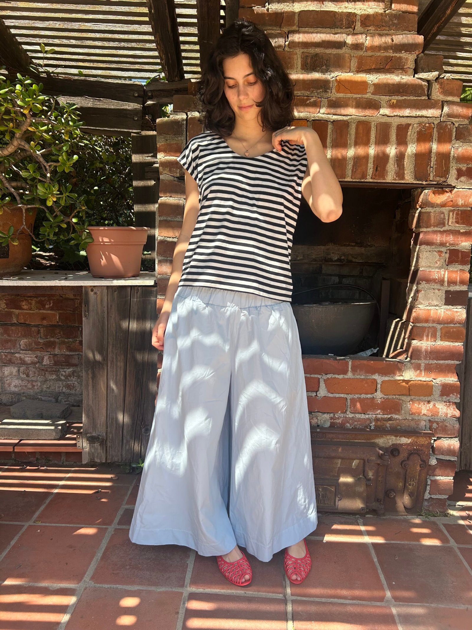 A woman stands in front of a brick fireplace outdoors, wearing a black and white striped shirt, light blue wide-leg pants, and pink sandals. Sunlight creates shadows on the ground.