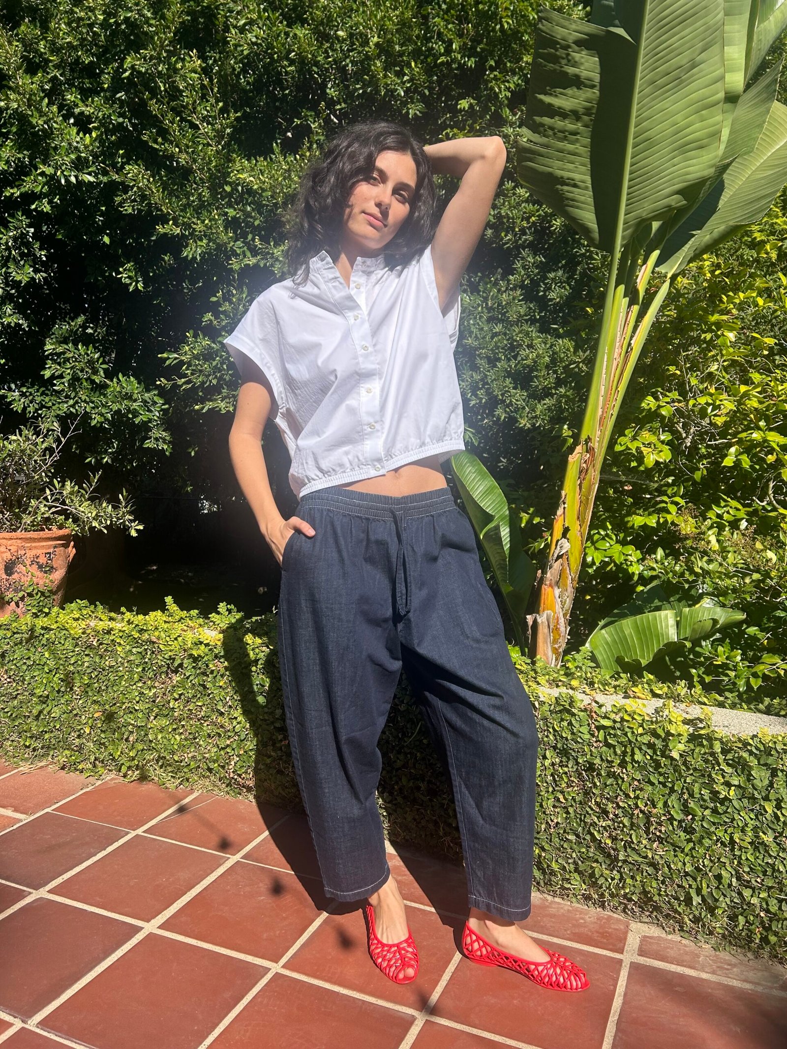 Woman standing on a tiled patio in front of greenery, wearing a white short-sleeve shirt, dark pants, and red shoes, with one hand raised behind her head.