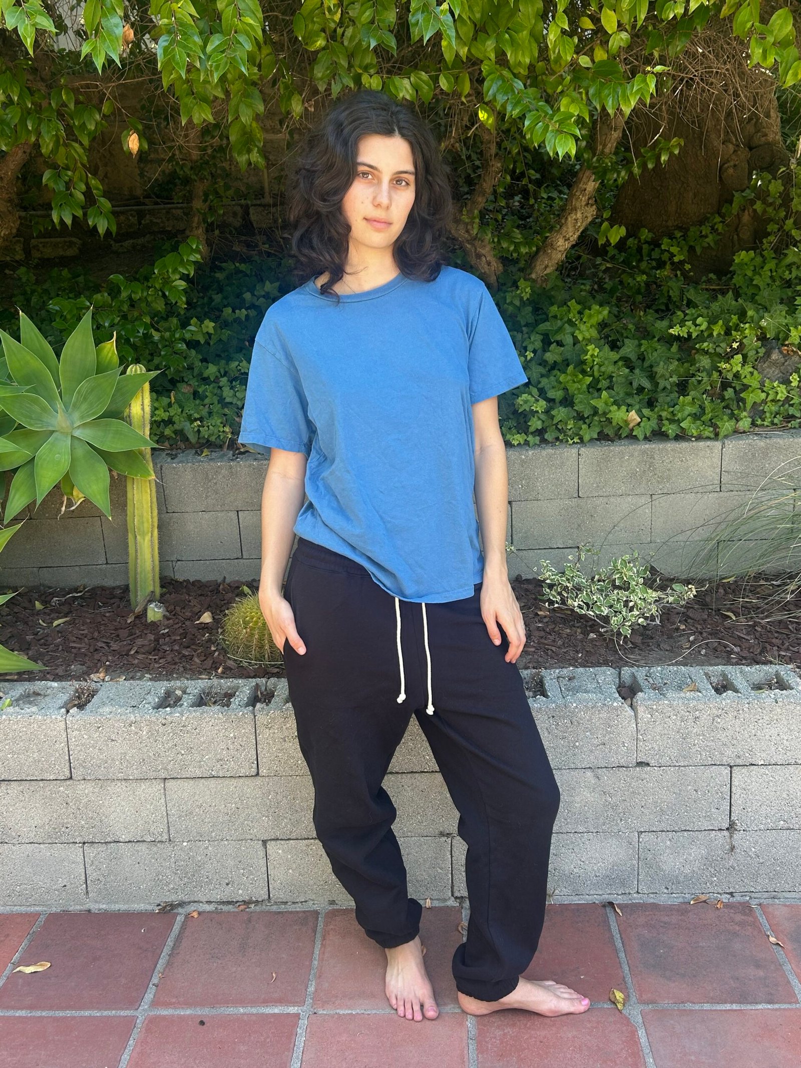 A person with long dark hair stands barefoot on a patio, wearing a blue t-shirt and black sweatpants, with greenery and a brick wall in the background.