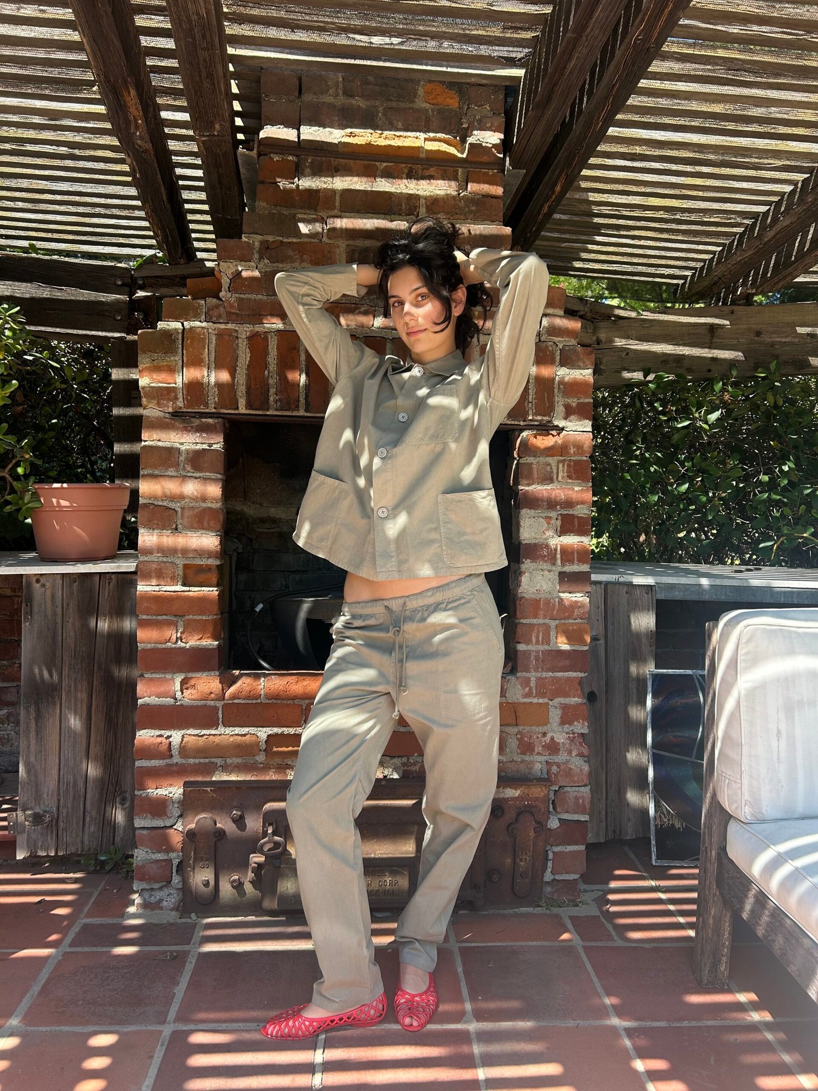 A person in a tan jacket and pants stands barefoot on a tiled patio in front of a brick outdoor fireplace, with sunlight filtering through a wooden pergola.