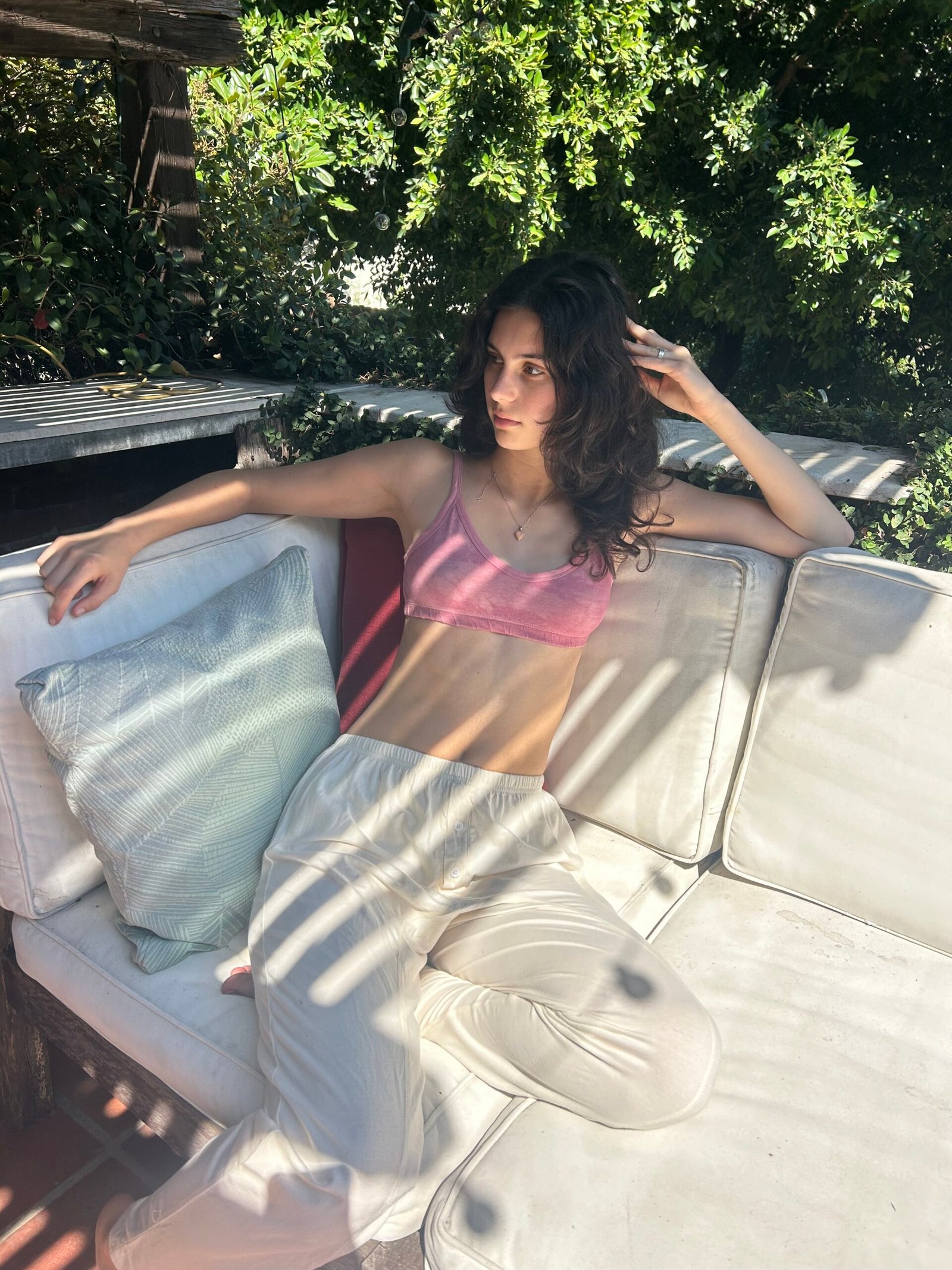 A woman in a pink crop top and white pants sits on a white outdoor sofa, resting her arm on a cushion, with sunlight and shadows from trees overhead.