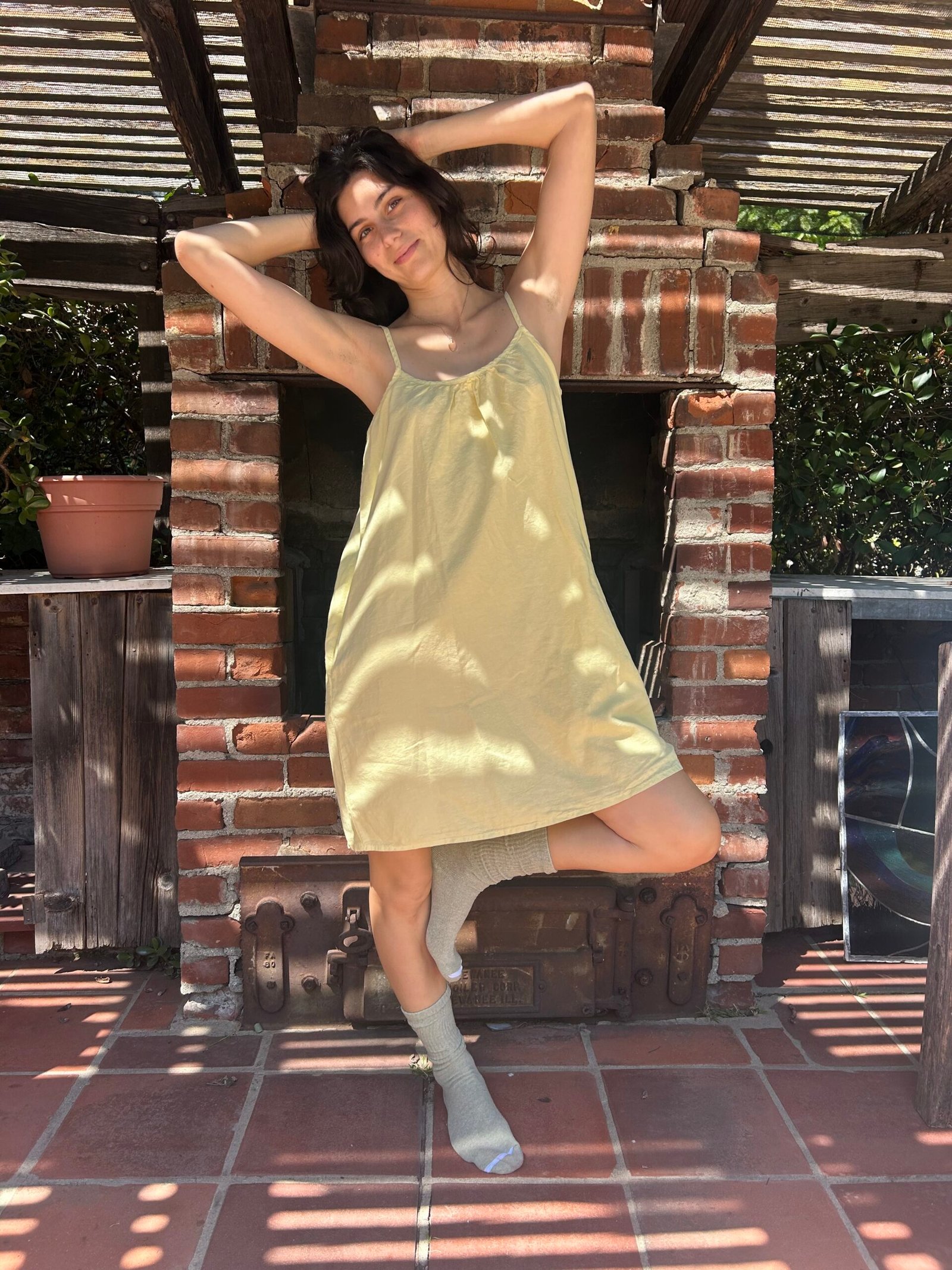 A woman in a light yellow dress and gray socks poses with arms raised and one leg bent in front of an outdoor brick fireplace under a pergola.