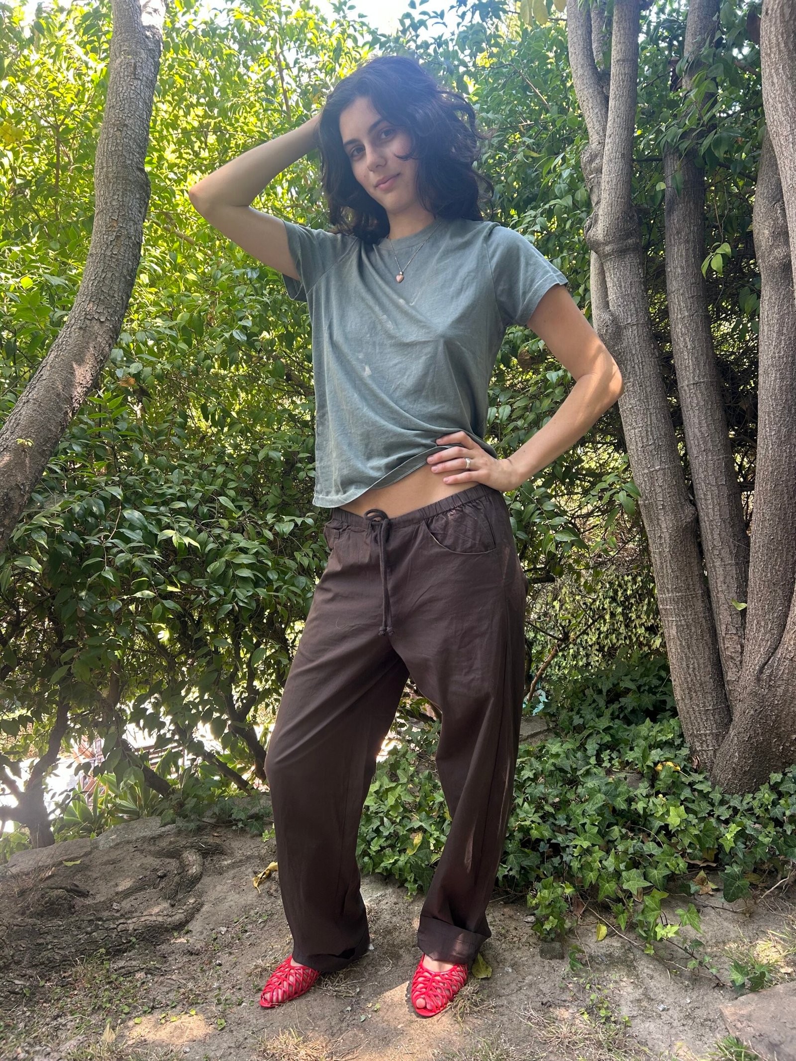 A person stands outdoors on a dirt path, wearing a green t-shirt, brown pants, and red shoes, with one hand on their head and the other on their hip, surrounded by trees and greenery.