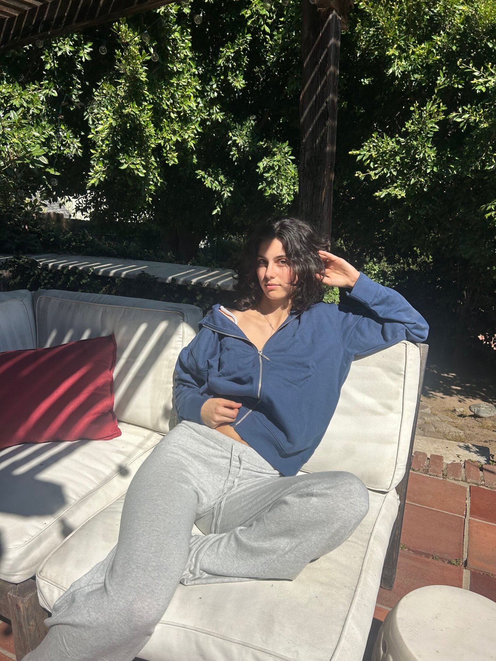 A person in a blue zip-up sweatshirt and gray sweatpants lounges on a white outdoor sofa under partial sunlight, with greenery in the background.
