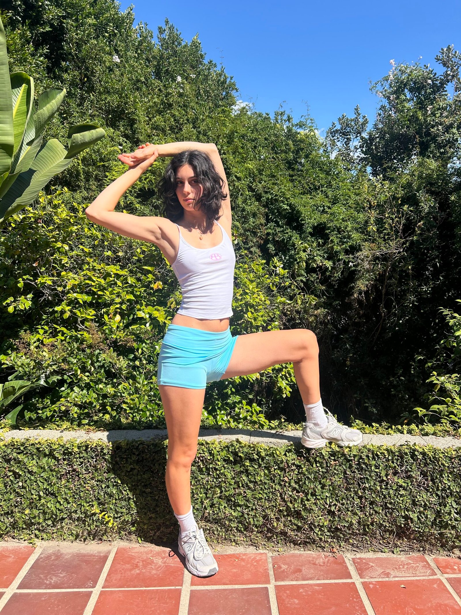 A person in a white tank top, blue shorts, and sneakers poses with one arm raised and one leg bent on a brick patio, with greenery and blue sky in the background.