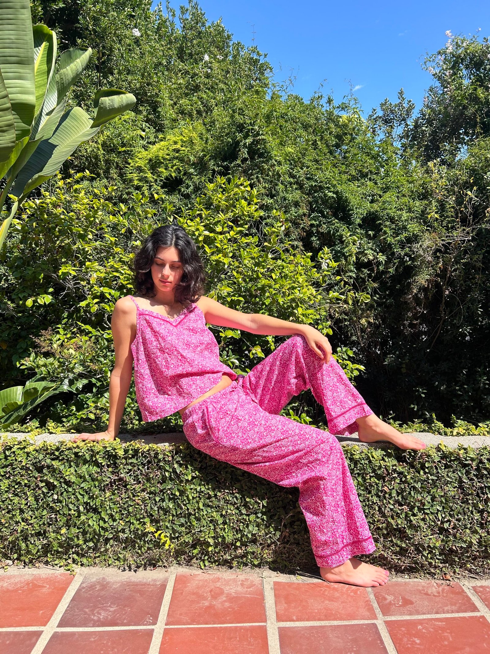A woman in pink floral pajamas sits on a low hedge in a garden with lush green foliage under a clear blue sky.