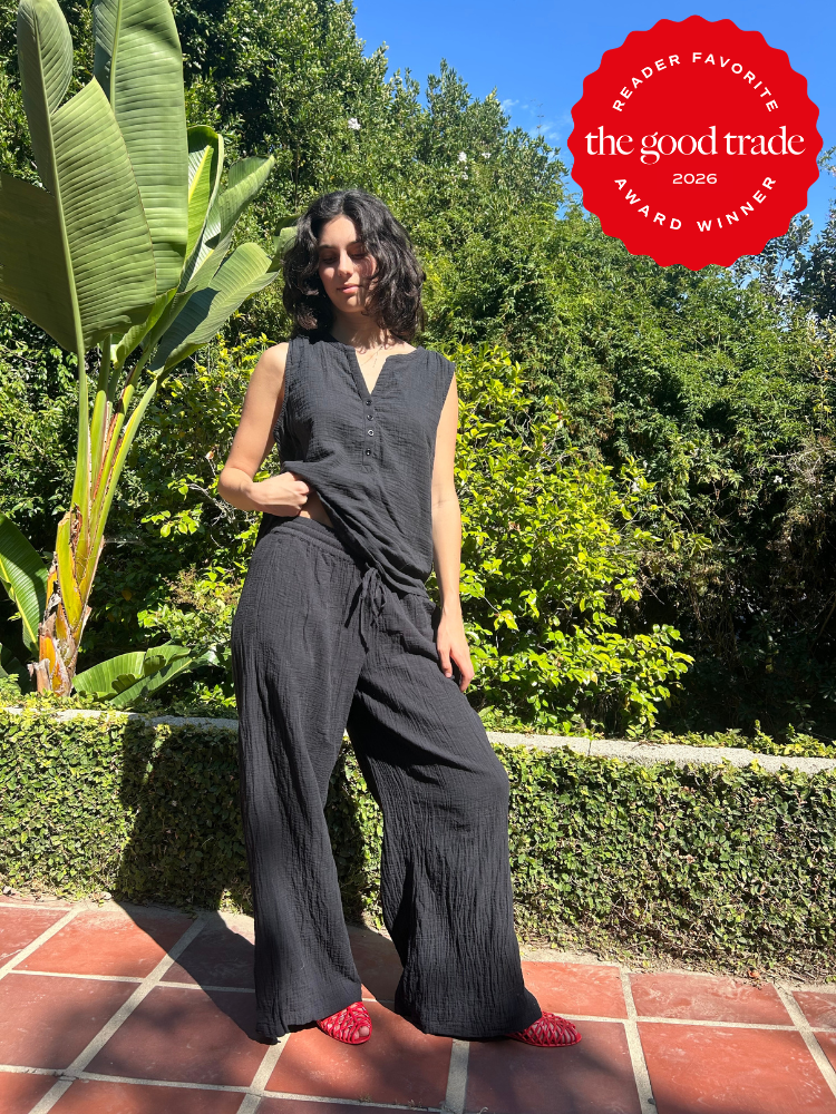 A woman in a loose black sleeveless jumpsuit and red shoes stands on a tiled patio with green plants in the background. A red "The Good Trade Award Winner 2026" badge is in the corner.