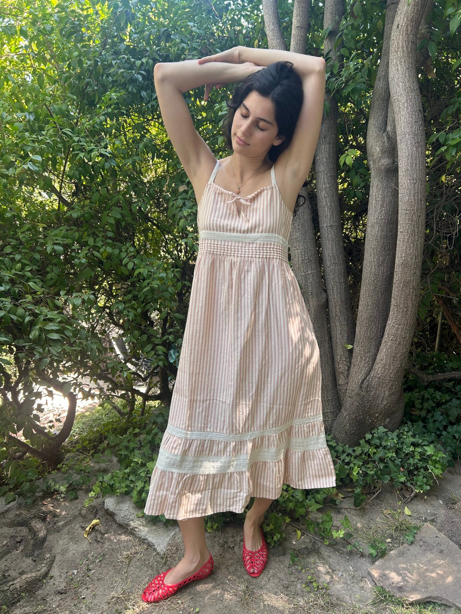Woman wearing a striped sundress and red shoes stands outdoors with her arms raised, surrounded by green foliage and trees.