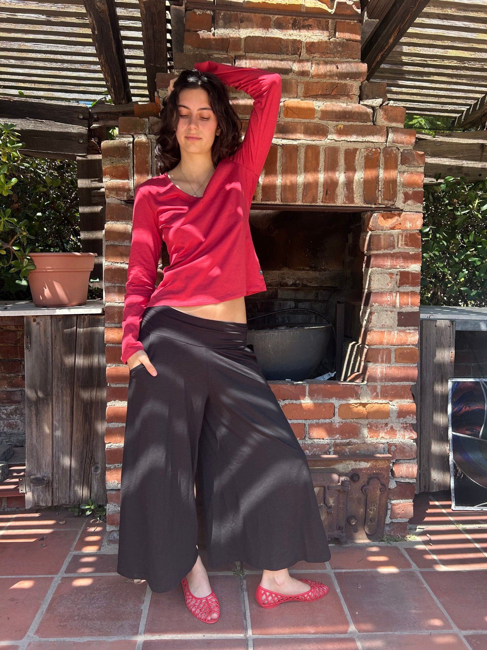 A woman stands in front of a brick outdoor fireplace, wearing a red long-sleeve top, black wide-leg pants, and red shoes, with one hand on her head and eyes closed.