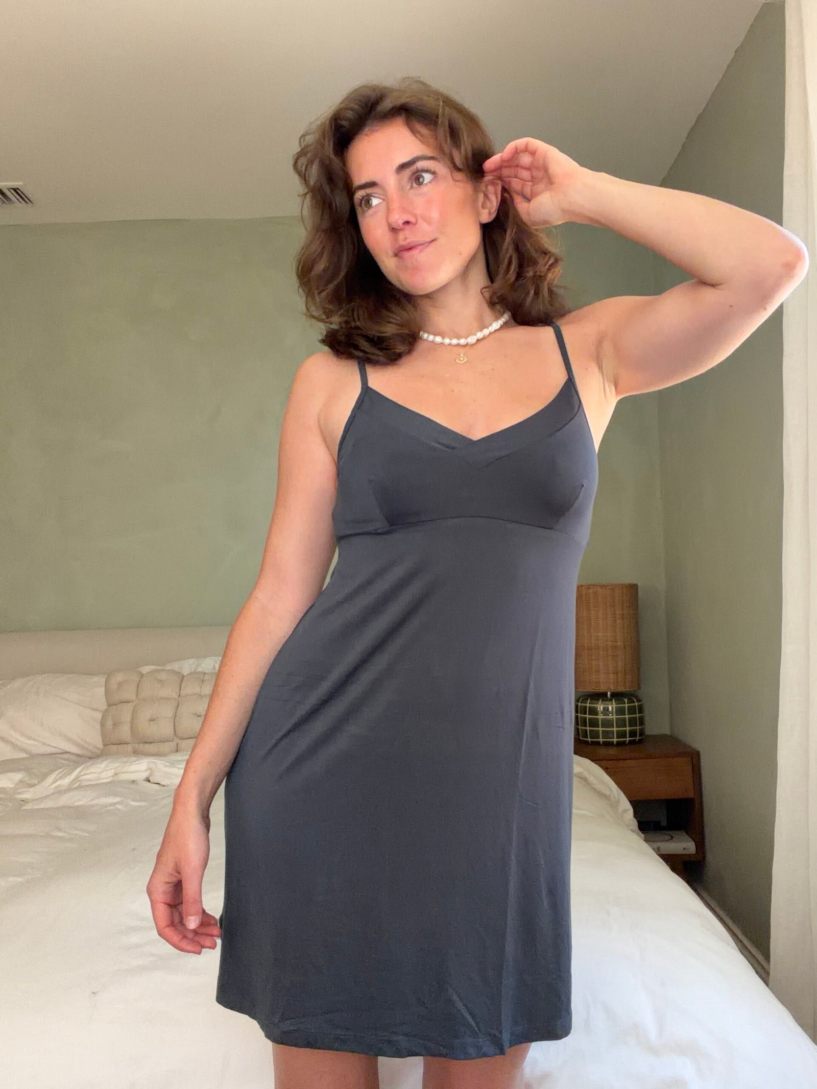 Woman stands in a bedroom wearing a dark slip dress and pearl necklace, one hand touching her ear, with an unmade bed and nightstand in the background.