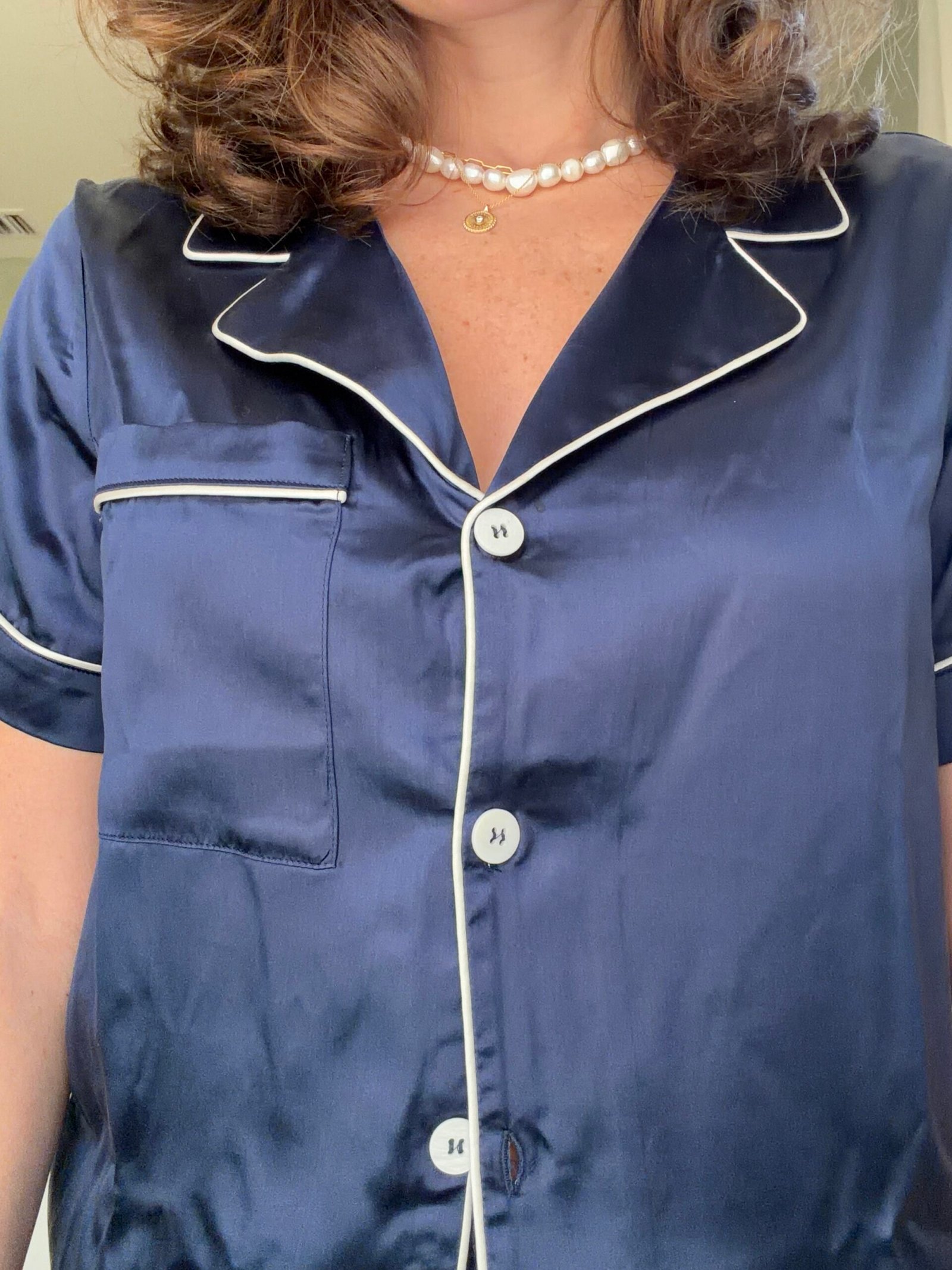 A person wearing a navy blue satin pajama top with white piping and a pearl necklace with a gold pendant.