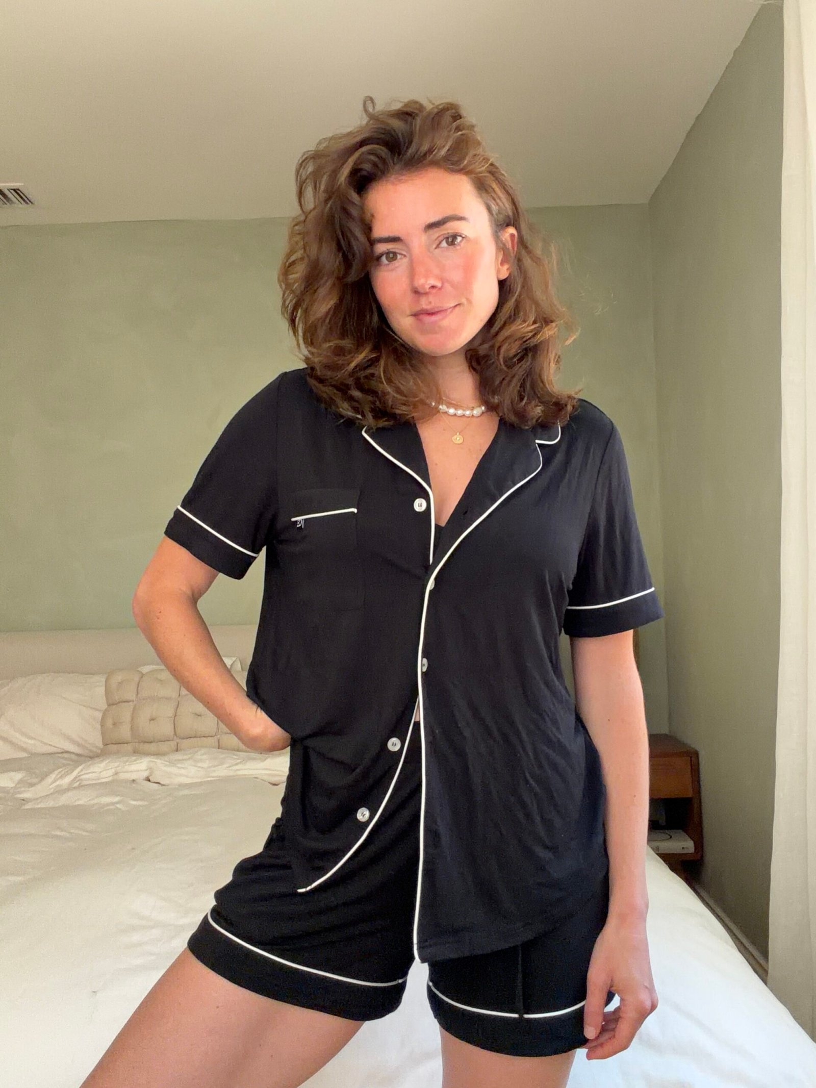 Woman stands indoors wearing a matching black pajama set with white trim, posing in front of an unmade bed in a simply furnished bedroom.
