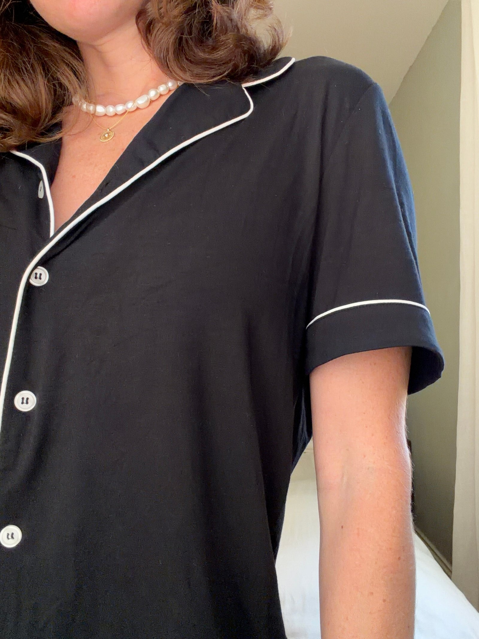 A person wearing a black pajama top with white piping and a pearl necklace, standing indoors near a bed.
