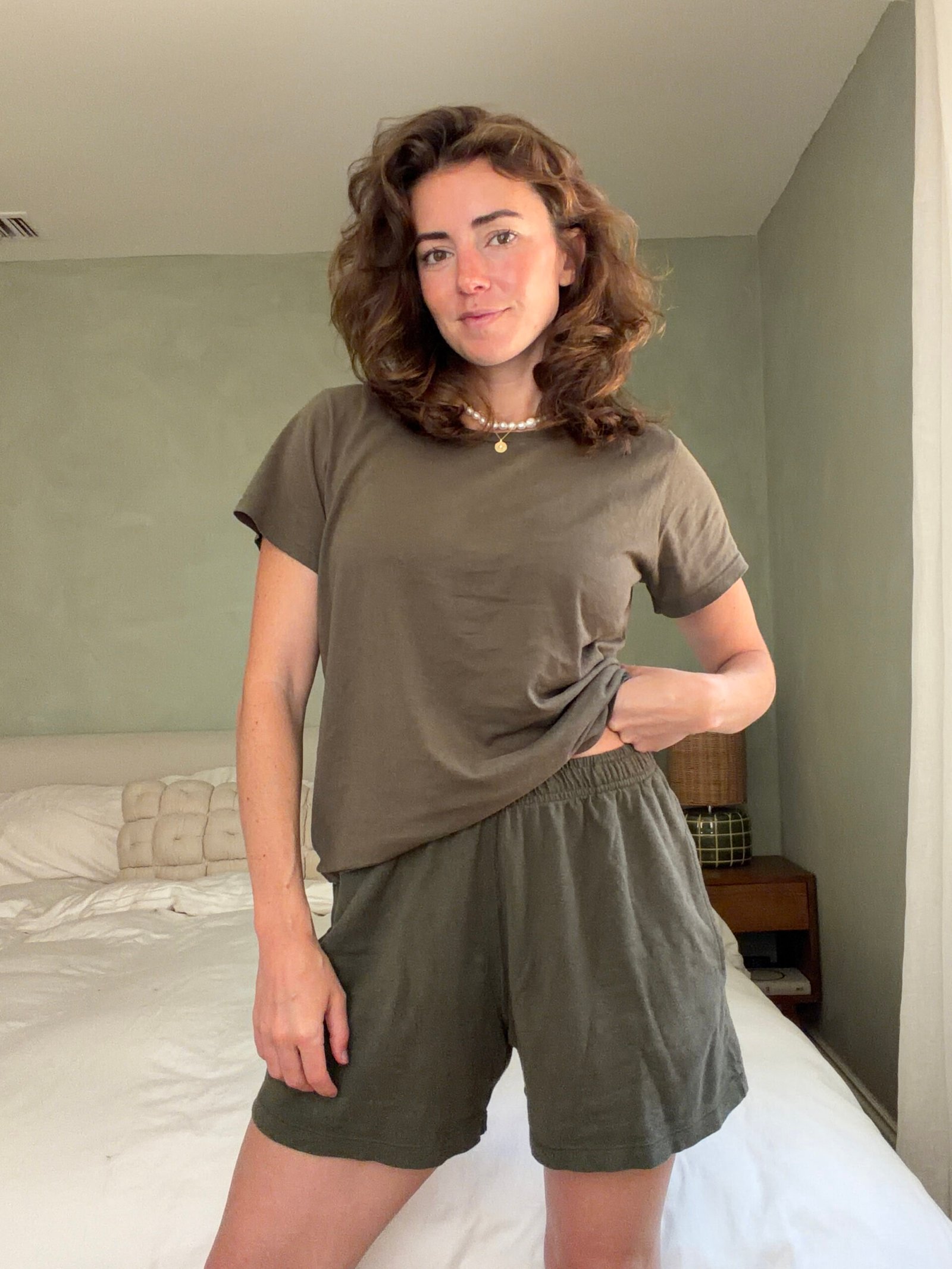 Woman standing in a bedroom wearing a brown T-shirt and green shorts, with one hand holding her shirt and the other by her side.