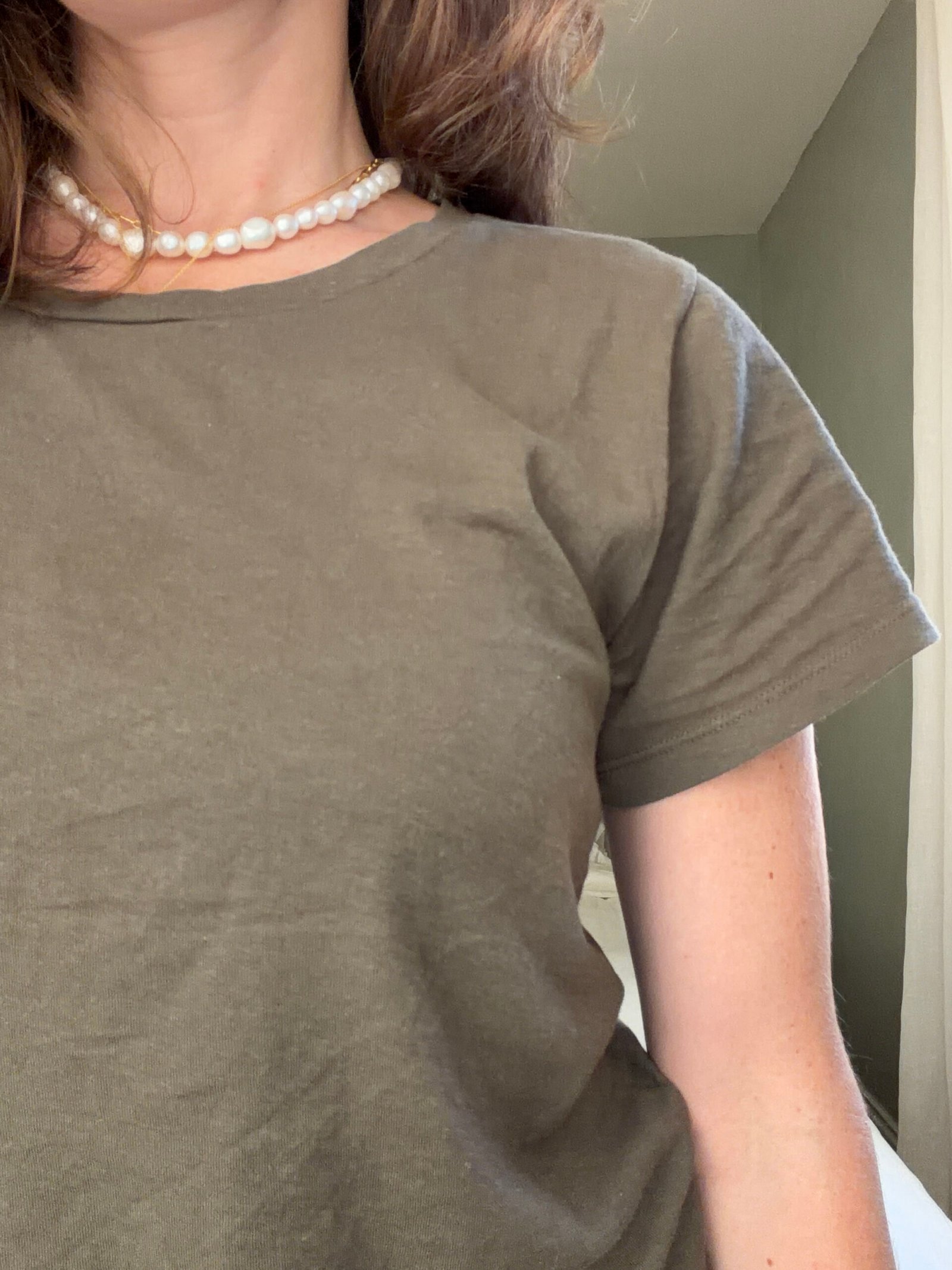 A person wearing a plain olive green t-shirt and a pearl necklace, shown from the shoulders to the upper chest, standing indoors.