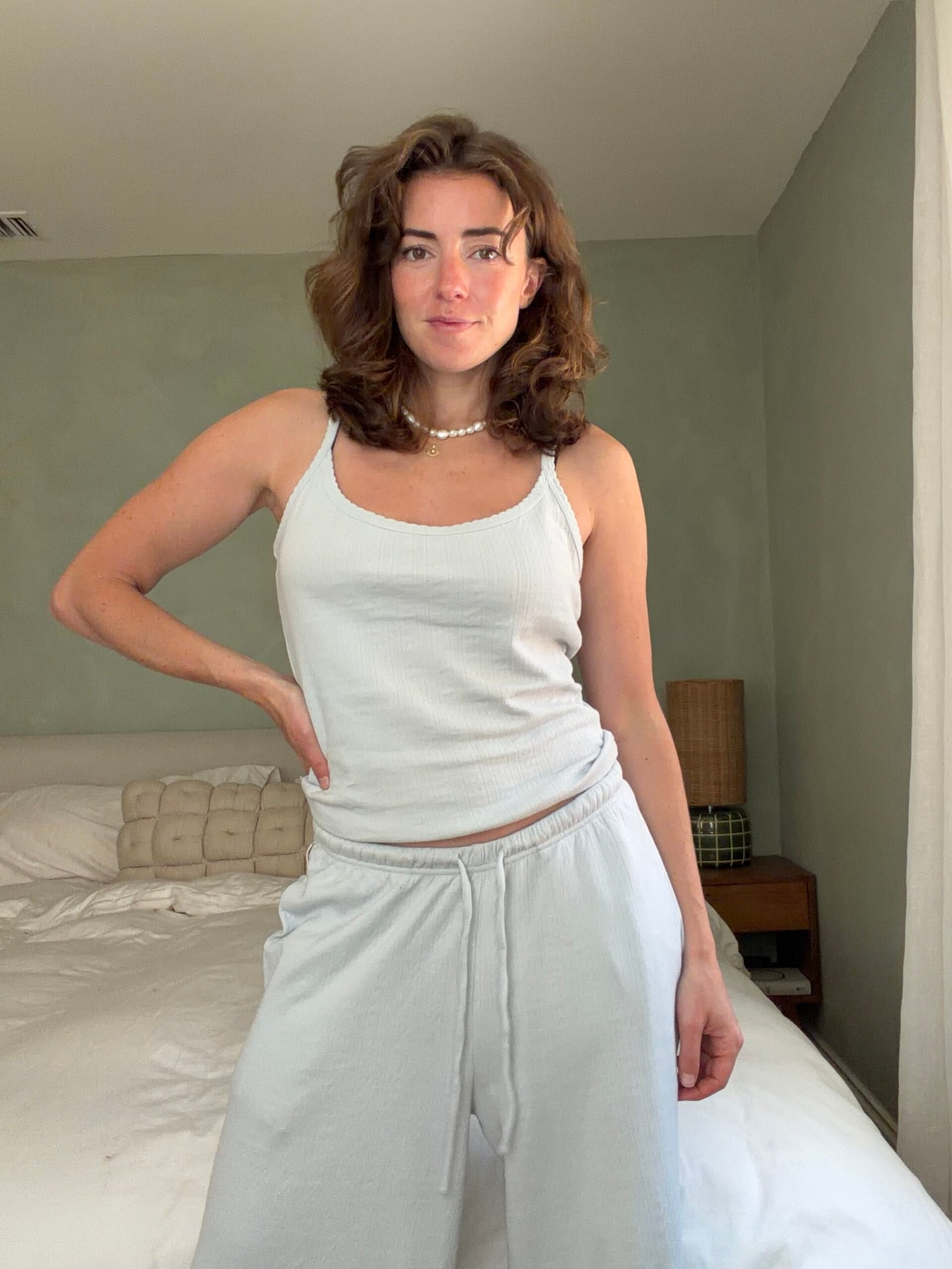 Woman stands in a bedroom wearing a white tank top and lounge pants, with one hand on her hip. A bed and side table are visible in the background.