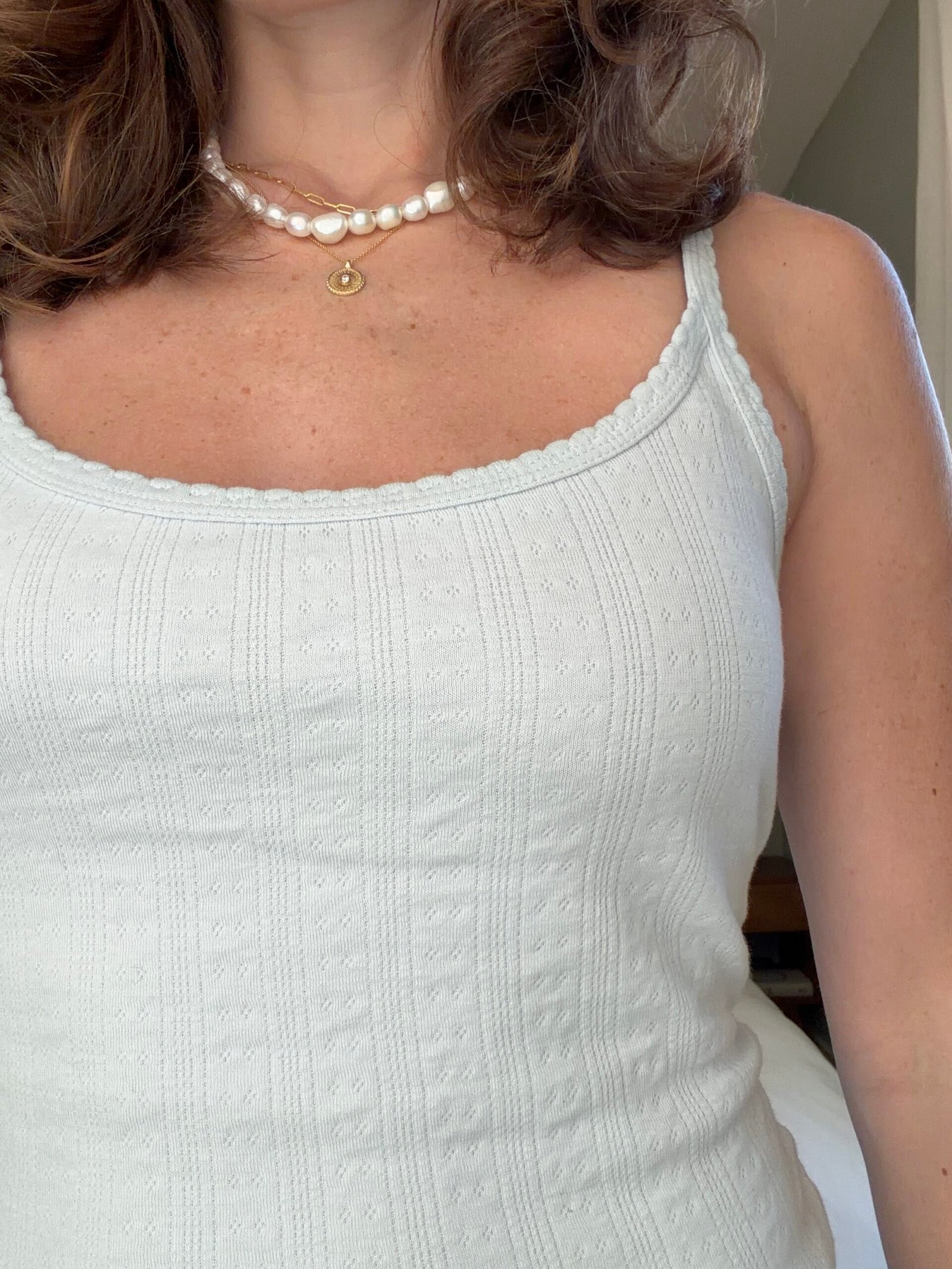 A woman wearing a white textured tank top, a pearl necklace, and a gold pendant necklace, shown from the shoulders to the waist.