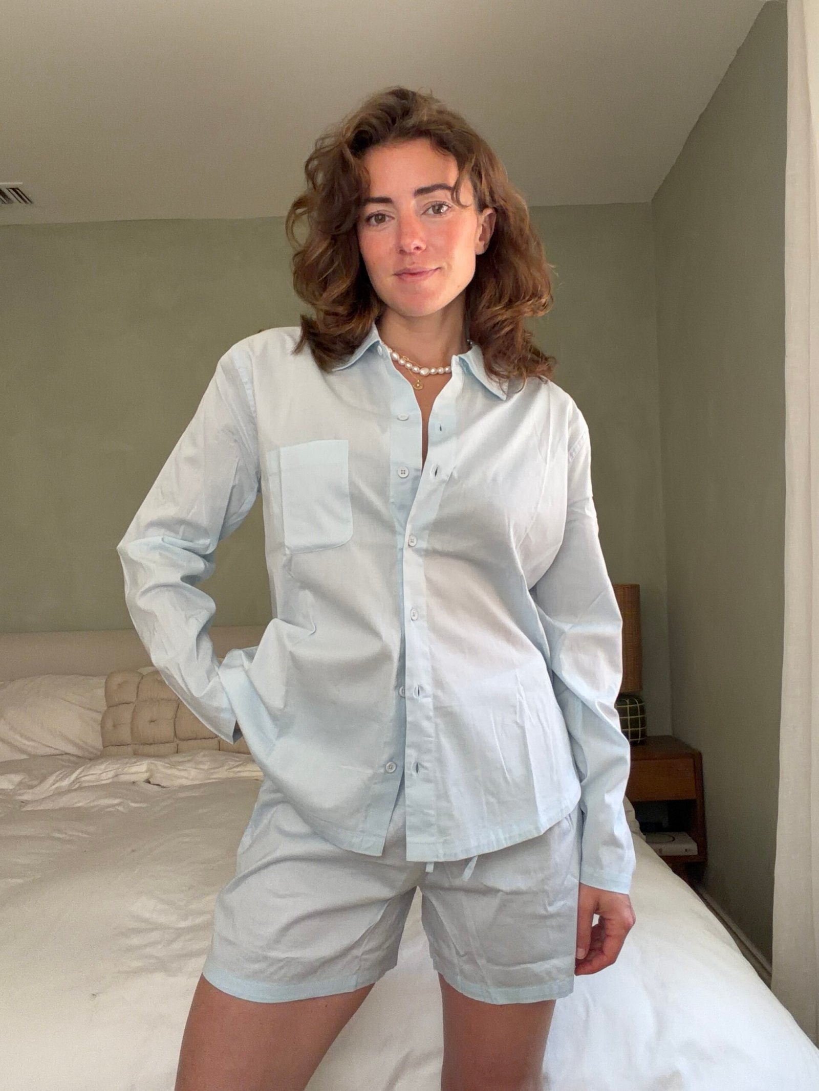 A person stands in a bedroom wearing a light blue button-up shirt and matching shorts, with one hand in their pocket, posing in front of an unmade bed.