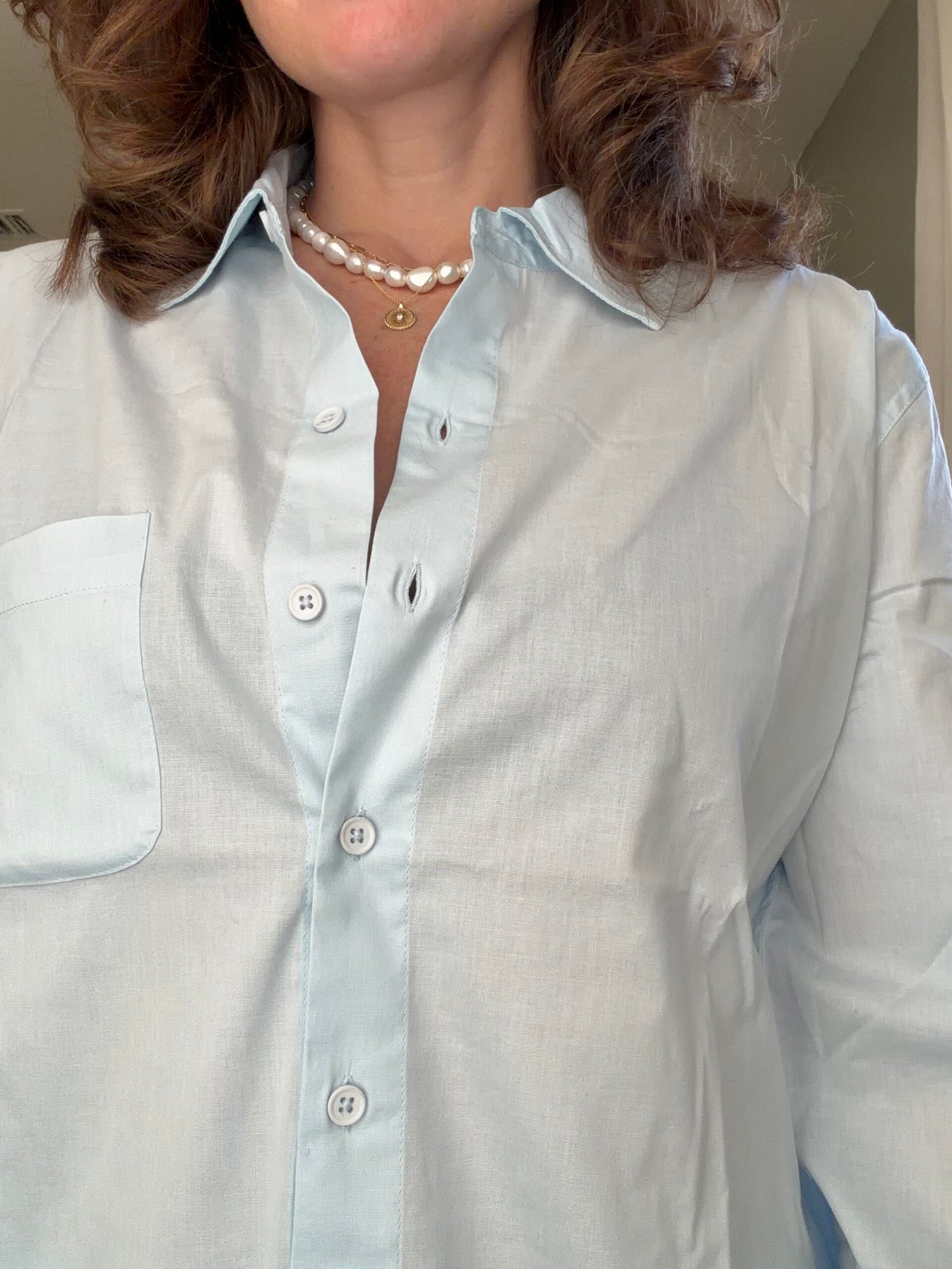 A woman wearing a light blue button-up shirt, a pearl necklace, and a gold pendant, shown from the shoulders to the mid-torso.