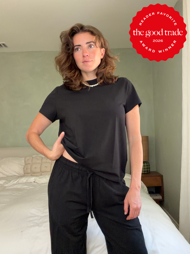 Woman stands in a bedroom wearing a black t-shirt and black pants. A red "The Good Trade Award Winner 2026" badge is in the top right corner.