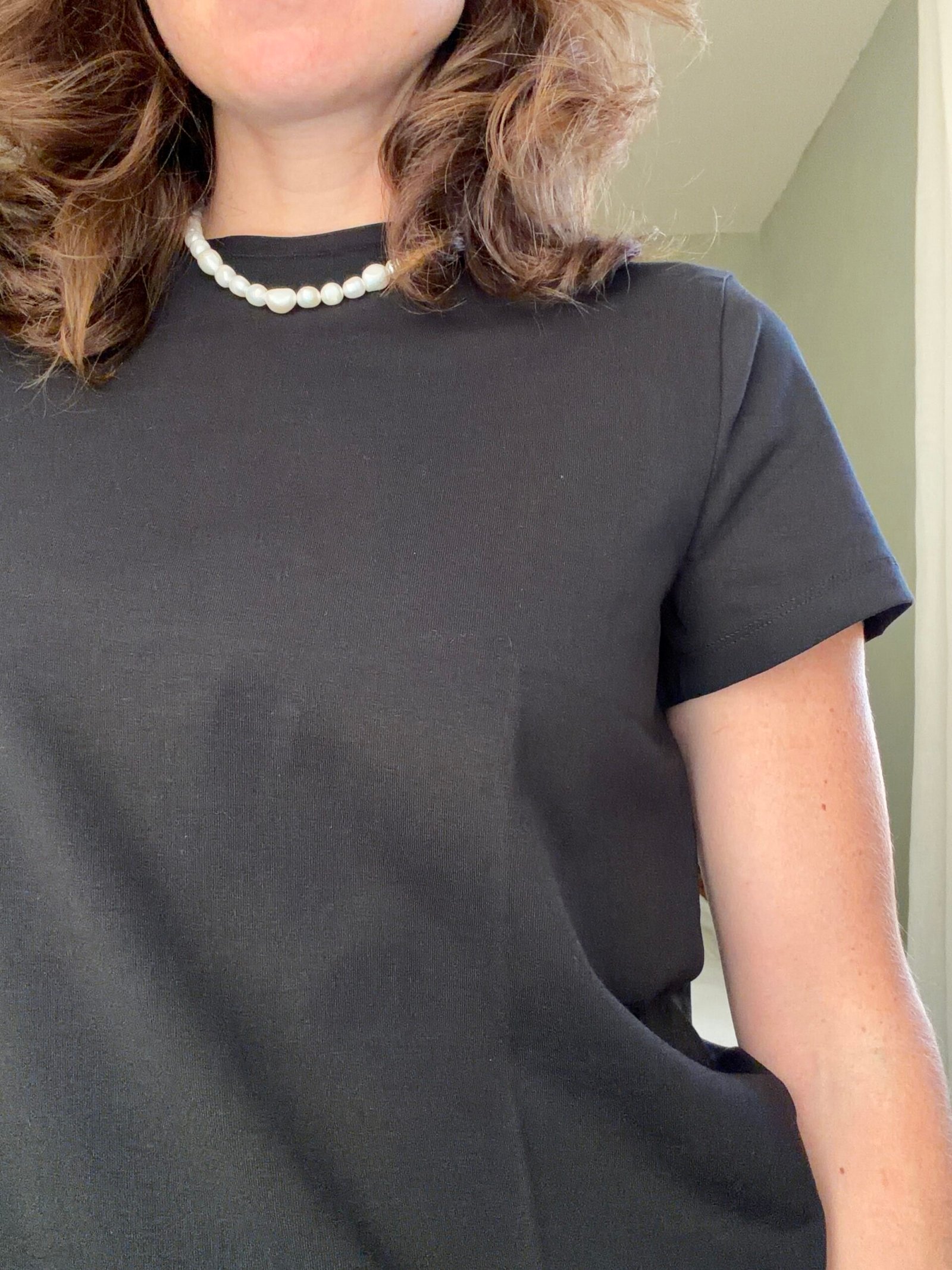 Person wearing a black short-sleeve top and a pearl necklace, shown from the shoulders to mid-torso. Face is partially visible.