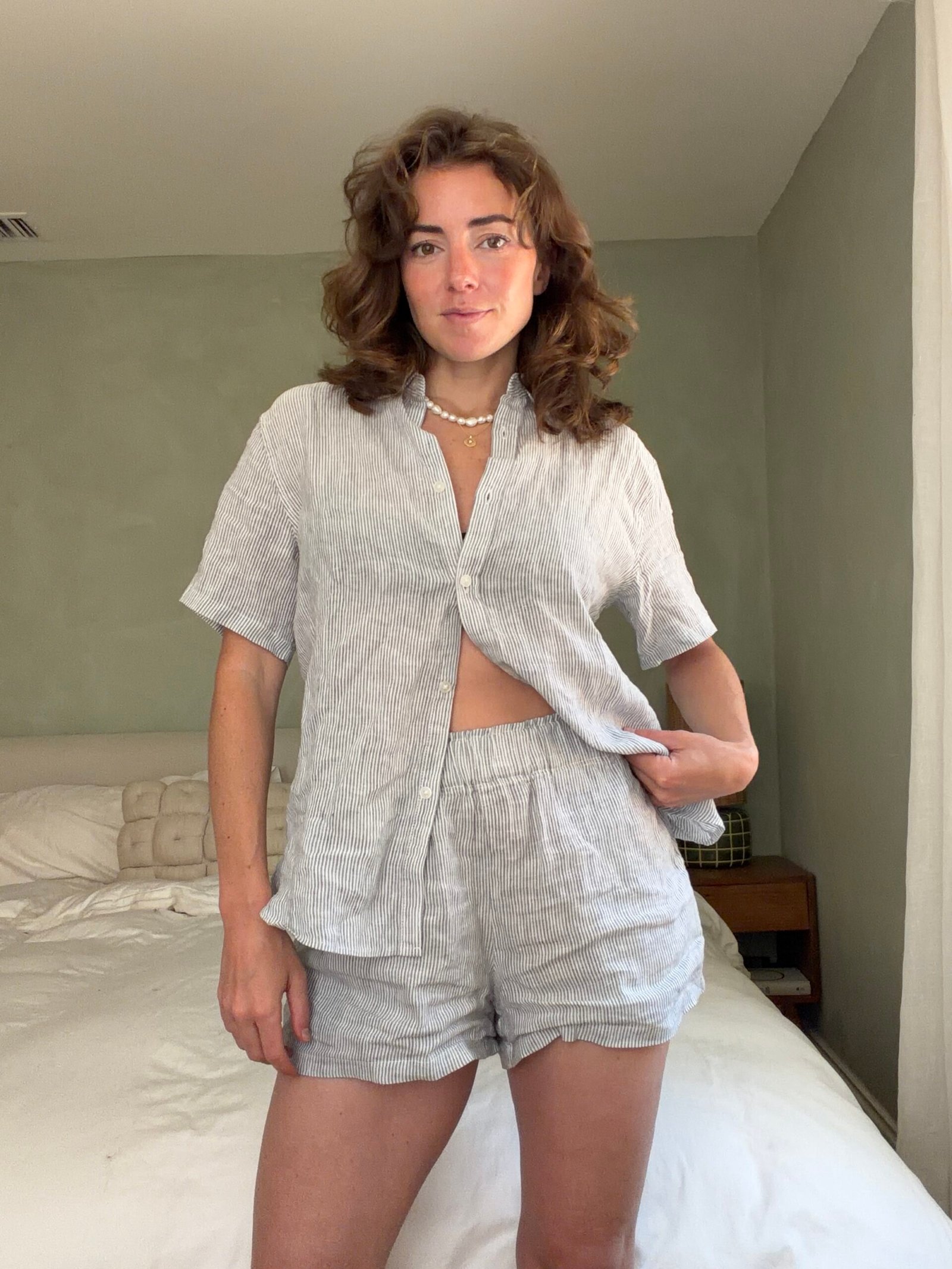 Woman stands in a bedroom wearing a light-colored, short-sleeve button-up shirt and matching shorts, with one hand holding her shirt open slightly.