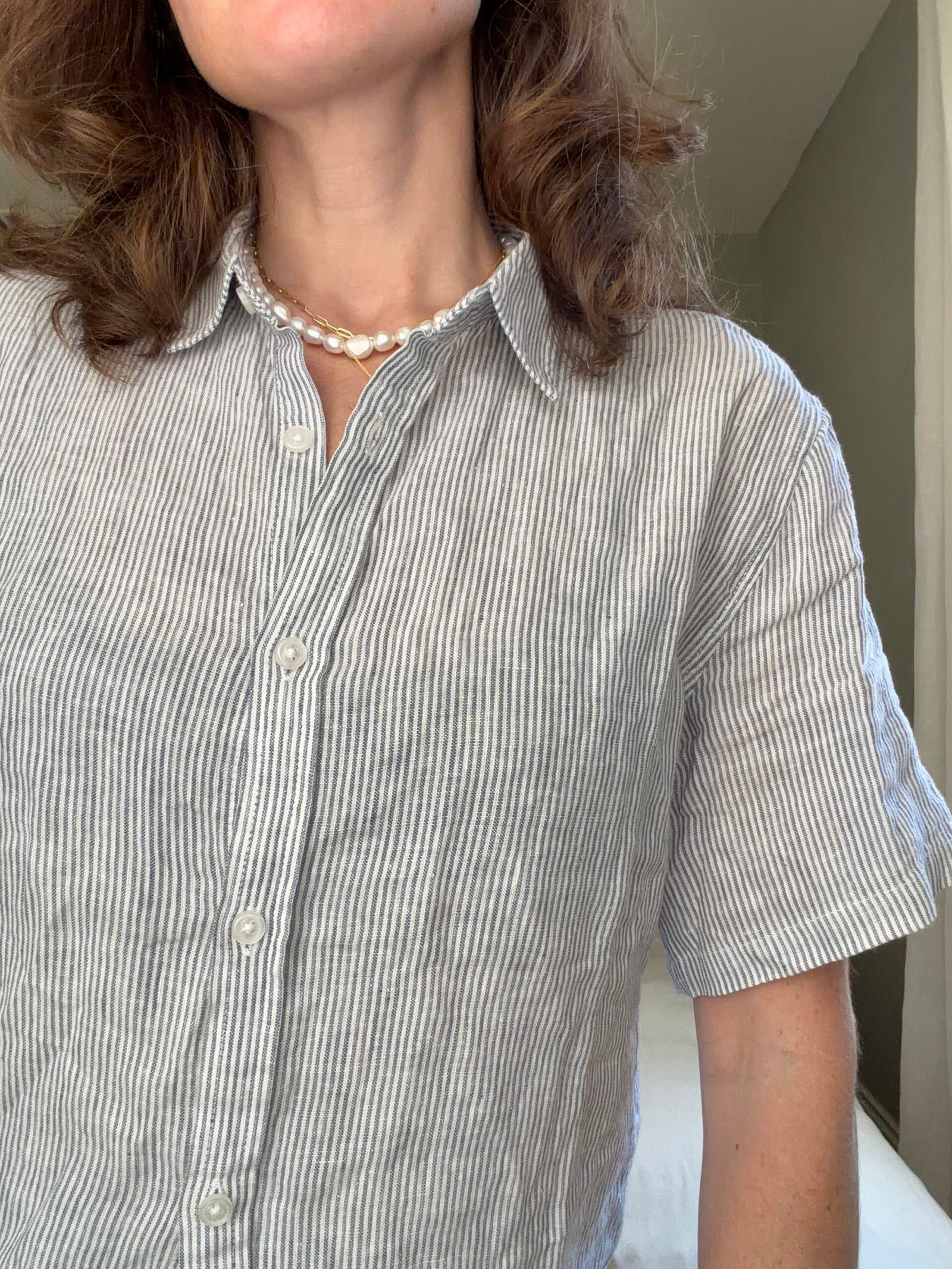Person wearing a short-sleeve, gray-and-white striped button-up shirt and a pearl necklace, standing indoors.