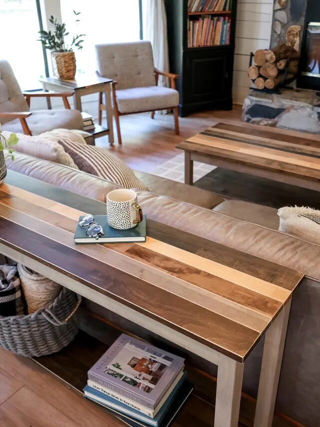 A living room with a sofa, two armchairs, a fireplace, and wooden tables. A striped console table behind the sofa holds a plant, books, cup, and other small items.