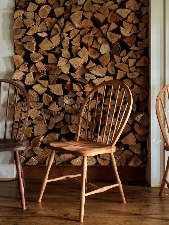 Three wooden chairs are placed in front of a stacked firewood wall on a wooden floor.