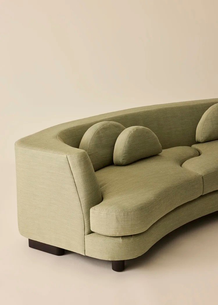 A curved, light green sofa with matching rounded cushions and dark wooden legs, set against a plain beige background.