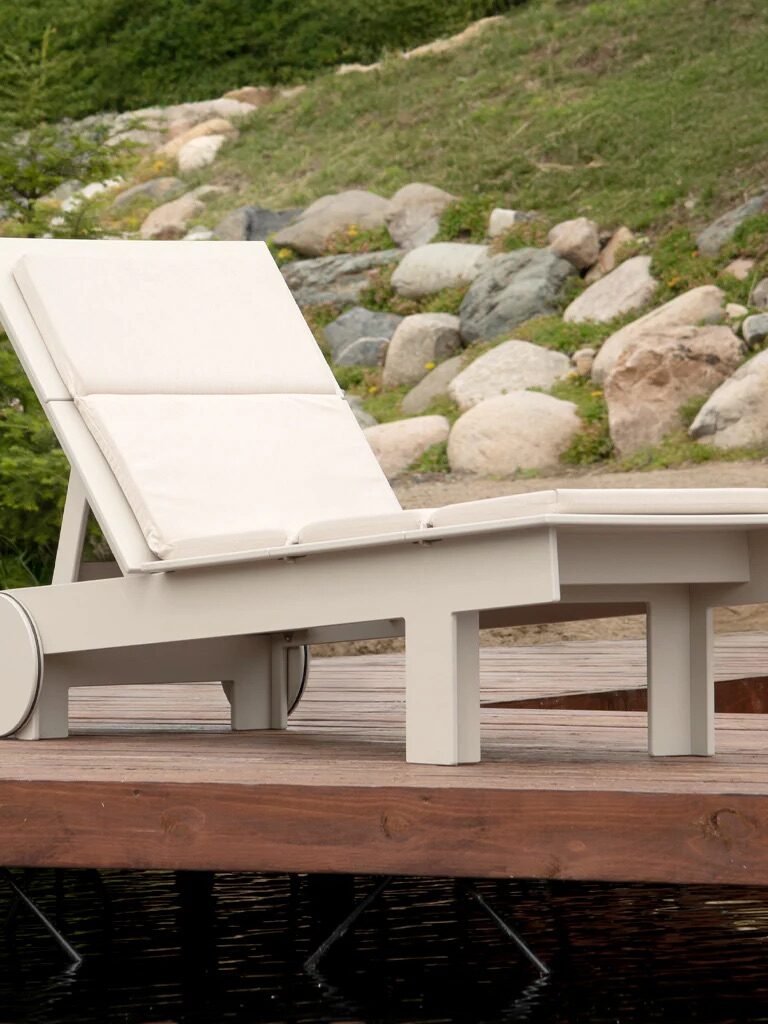 A white cushioned lounge chair with wheels is placed on a wooden deck near a grassy area and rocks.