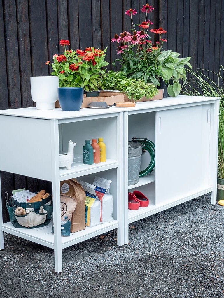 A white outdoor storage bench holds potted plants, gardening tools, bags of soil, watering cans, plant food, gloves, small boots, and pots, set against a dark wooden fence.