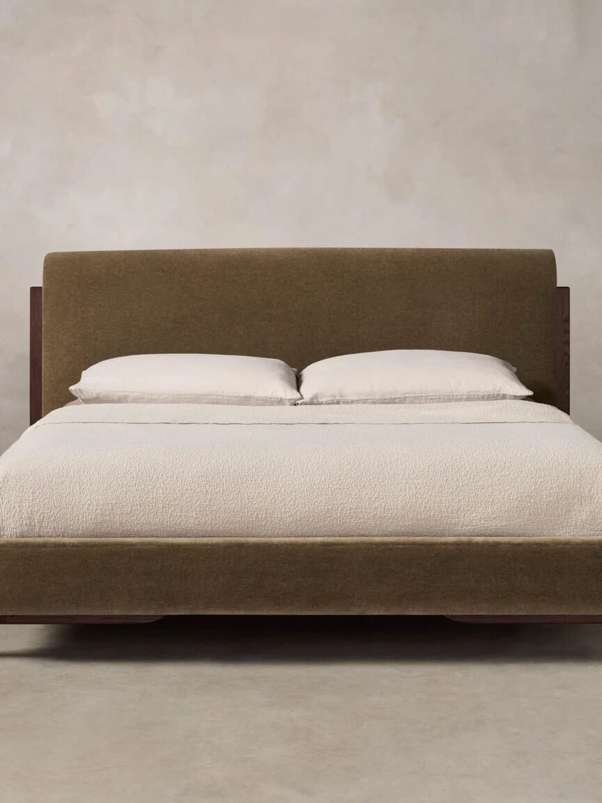 A modern bed with a brown upholstered frame and headboard, neatly made with white bedding and two pillows, against a plain light gray background.