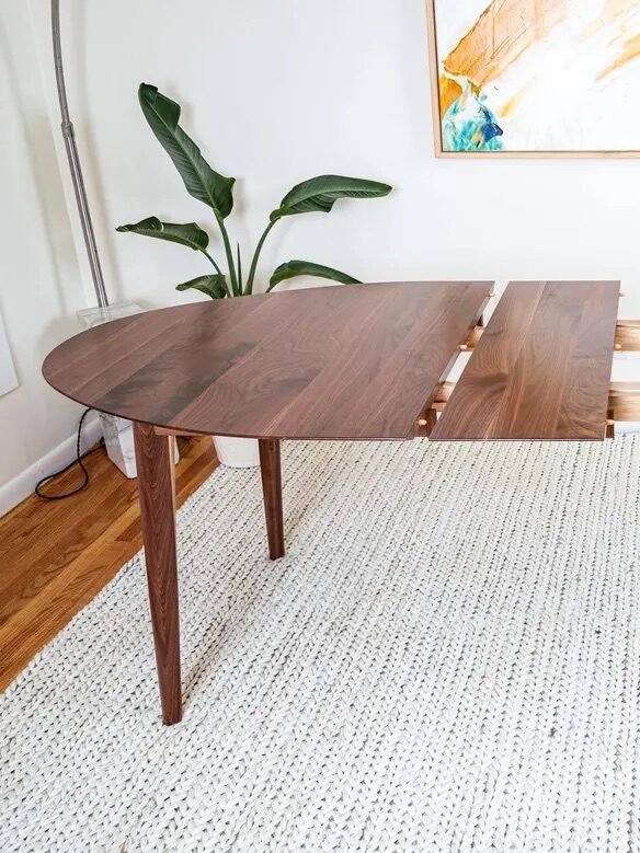 A wooden dining table with an extension mechanism partially opened, placed on a light-colored rug in a modern room with plants and abstract wall art.