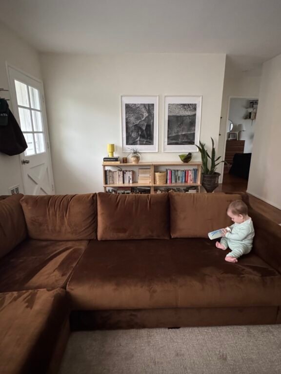 A baby sits on a brown sectional sofa holding a bottle in a living room with bookshelves, framed art, and a door in the background.