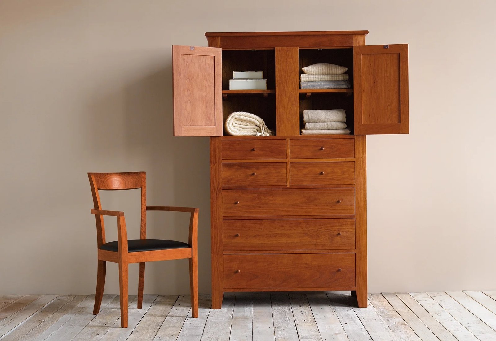 A wooden dresser with upper cabinet doors open revealing folded towels and linens, next to a matching wooden chair with a dark cushioned seat on a wooden floor.