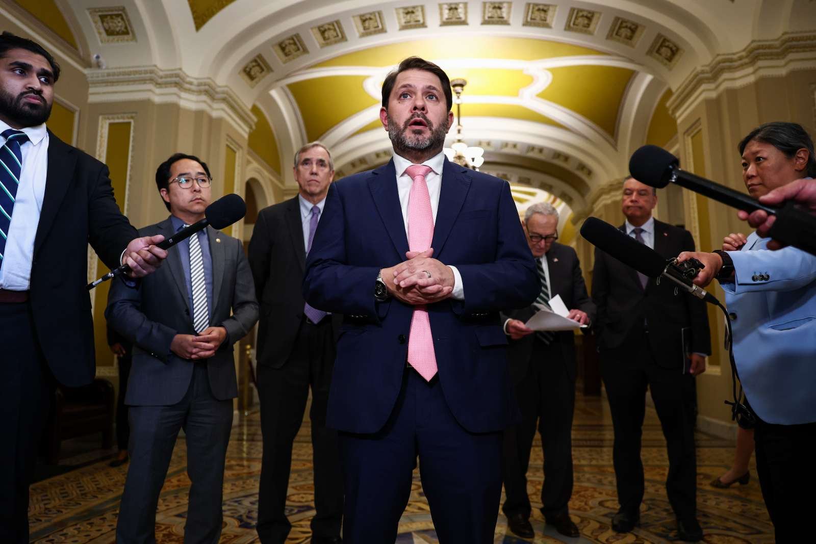 A man in a suit speaks in a government building surrounded by microphones