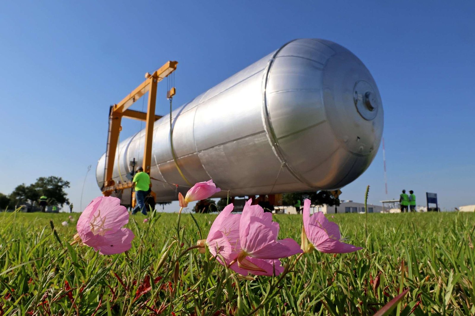 In the foreground, a few pink flowers on a grassy field lean to the right. Behind them is a long, silver cylinder being moved by crews in bright green safety shirts and vests. The massive object is the liquid hydrogen tank for the Artemis IV rocket.