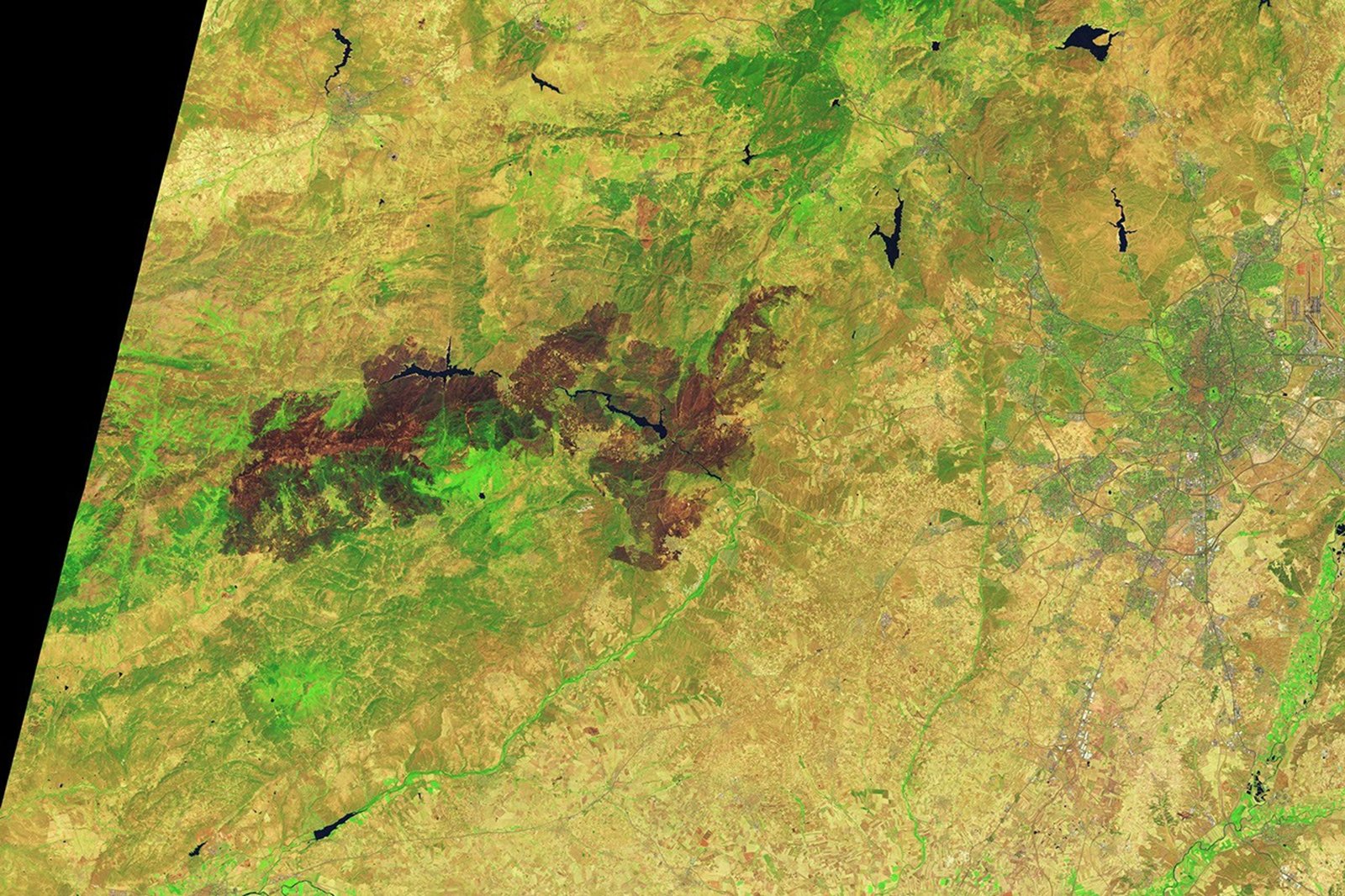 A brown patch of burned vegetation is visible on the landscape surrounding two reservoirs in a hilly region west of Madrid.