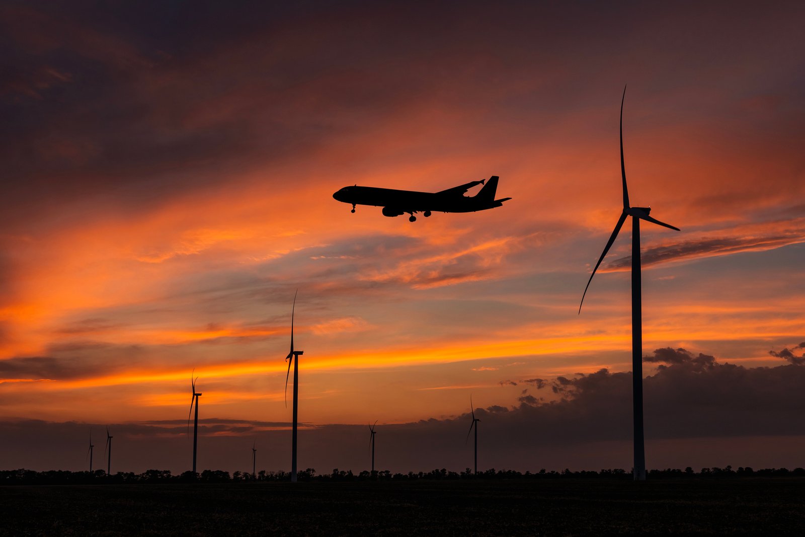 An airliner flies over a line of wind turbines at sunset.