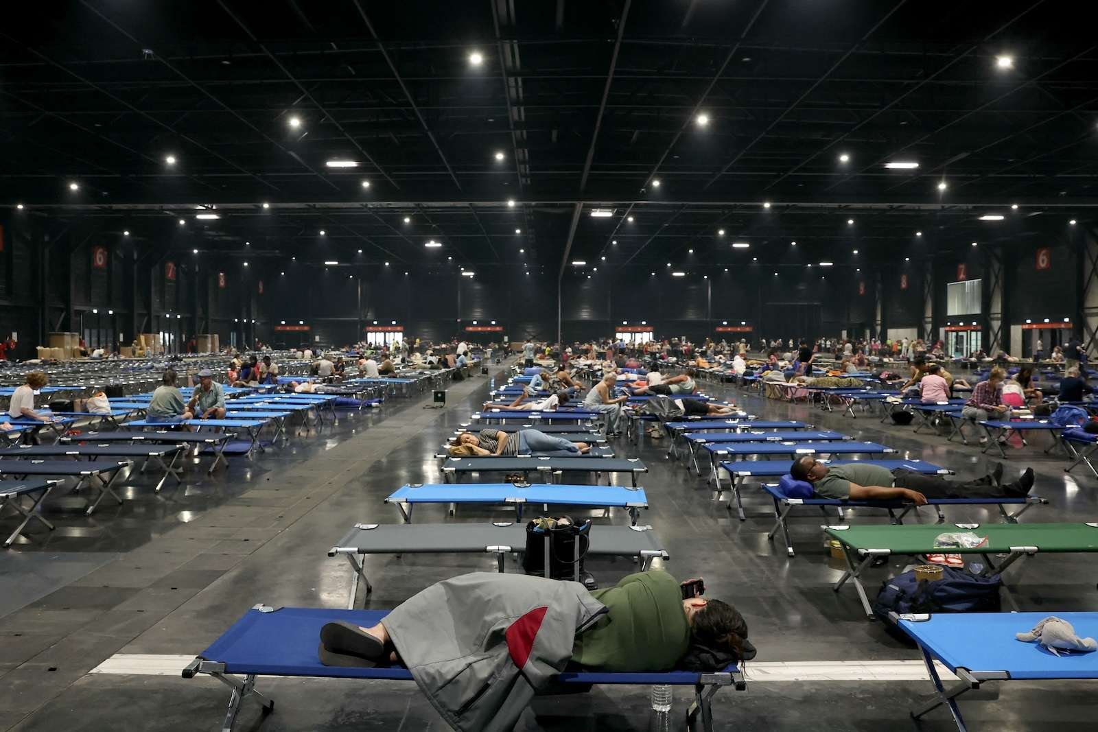 several rows of blue and grey cots, with dozens per row, strech into a large auditorium space. dozens of people are visible sitting, lounging, or sleeping on them.