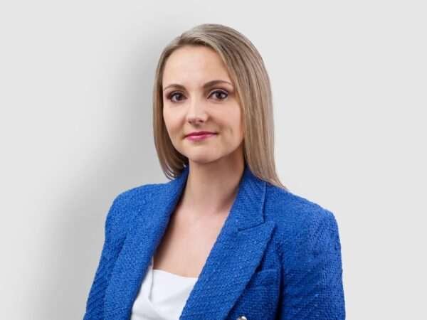 A woman with straight, light brown hair, wearing a textured blue blazer over a white top, stands against a neutral background. 